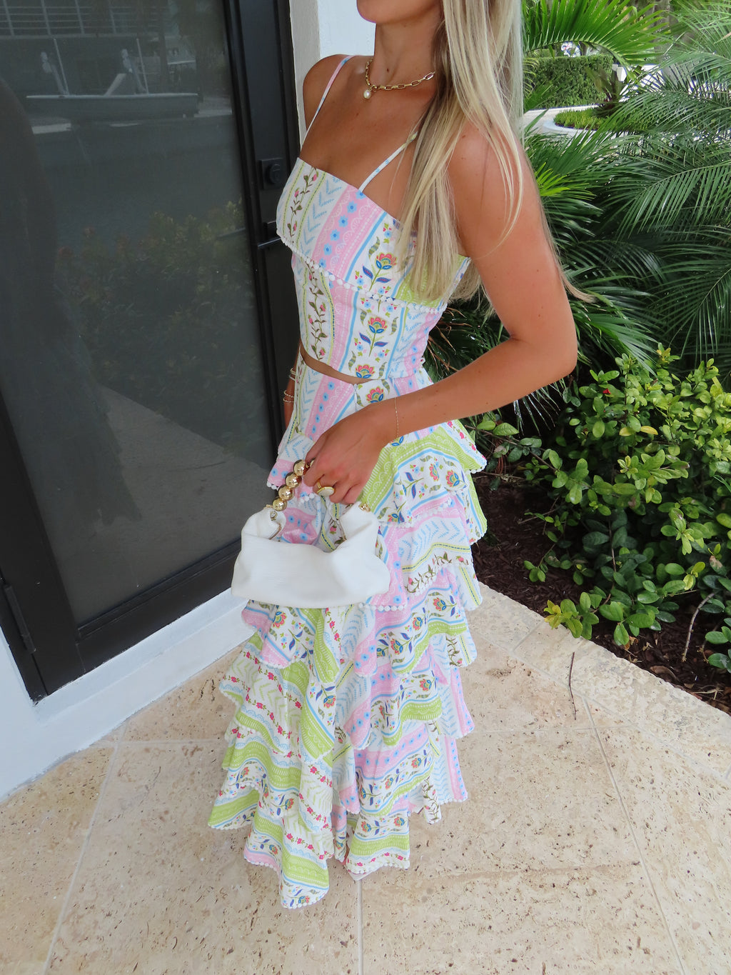 Woman holding a white purse in a patterned maxi skirt set standing near a glass door with greenery outside.