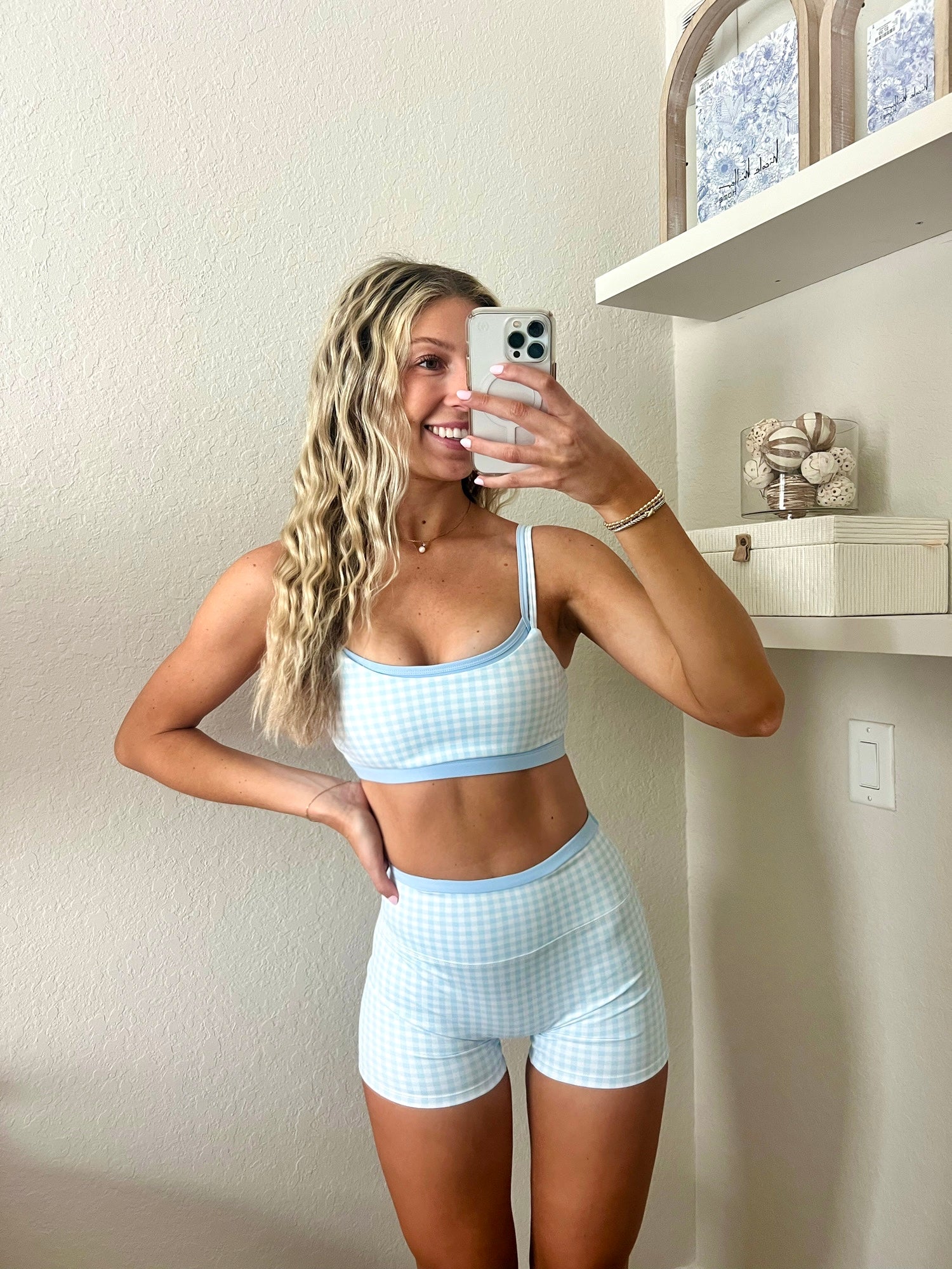 Woman taking a mirror selfie wearing a light blue and white gingham workout set, consisting of a sports bra and biker shorts, in a home setting.