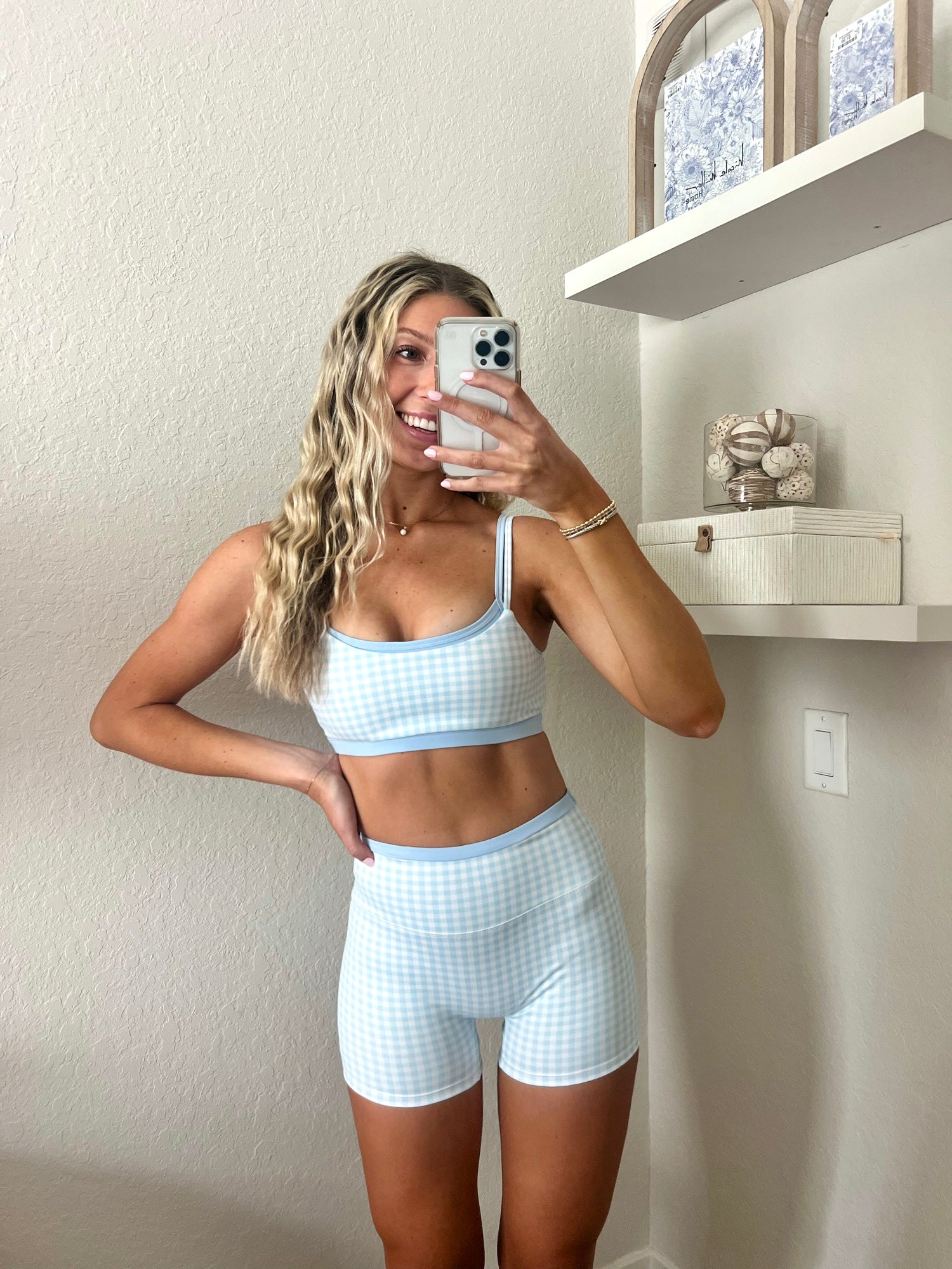 Woman taking a mirror selfie wearing a light blue and white gingham workout set, consisting of a sports bra and biker shorts, in a home setting.