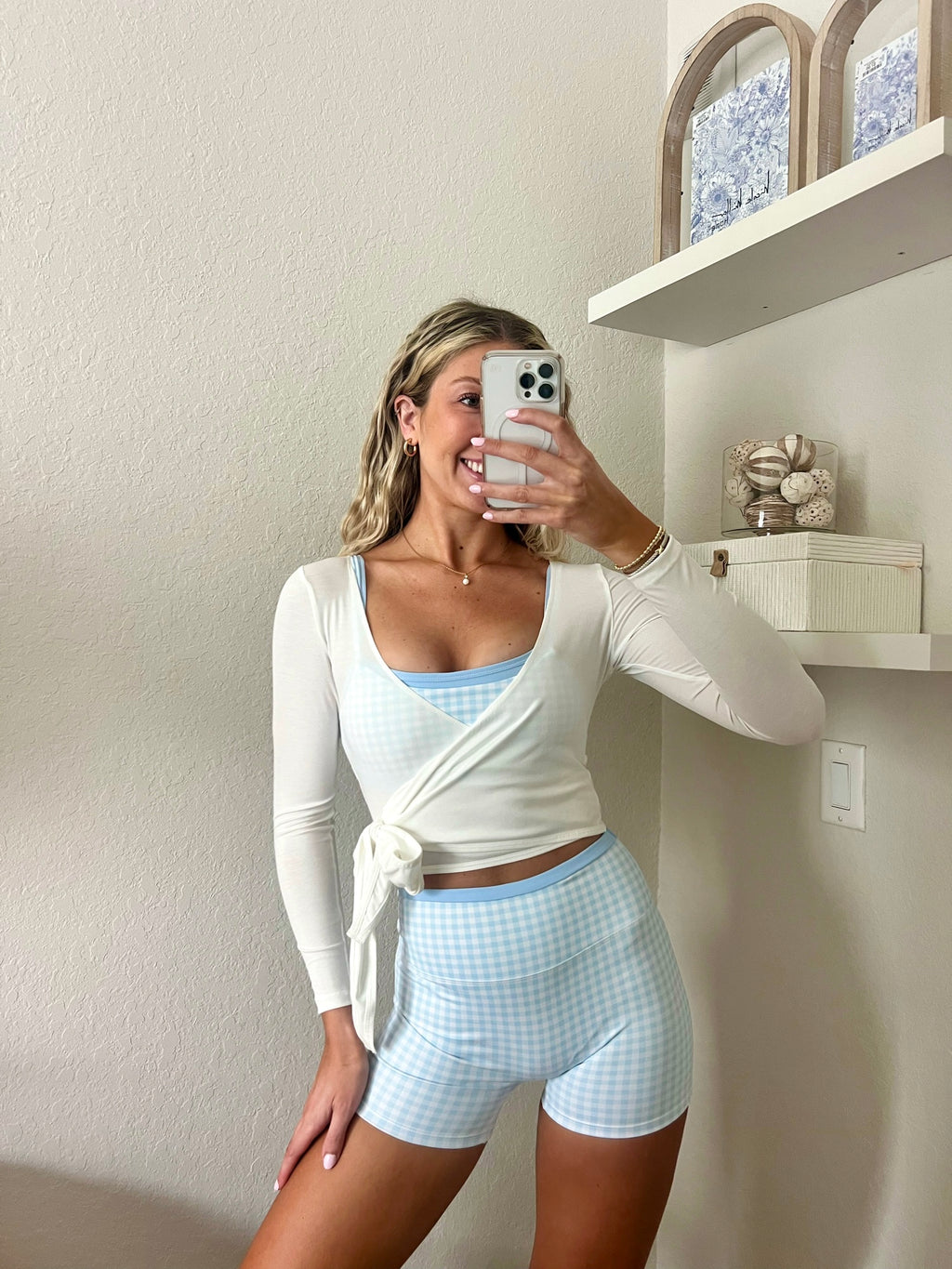 Woman taking a mirror selfie wearing a light blue and white gingham workout set, consisting of a sports bra and biker shorts, with a white long sleeve wrap top, in a home setting.