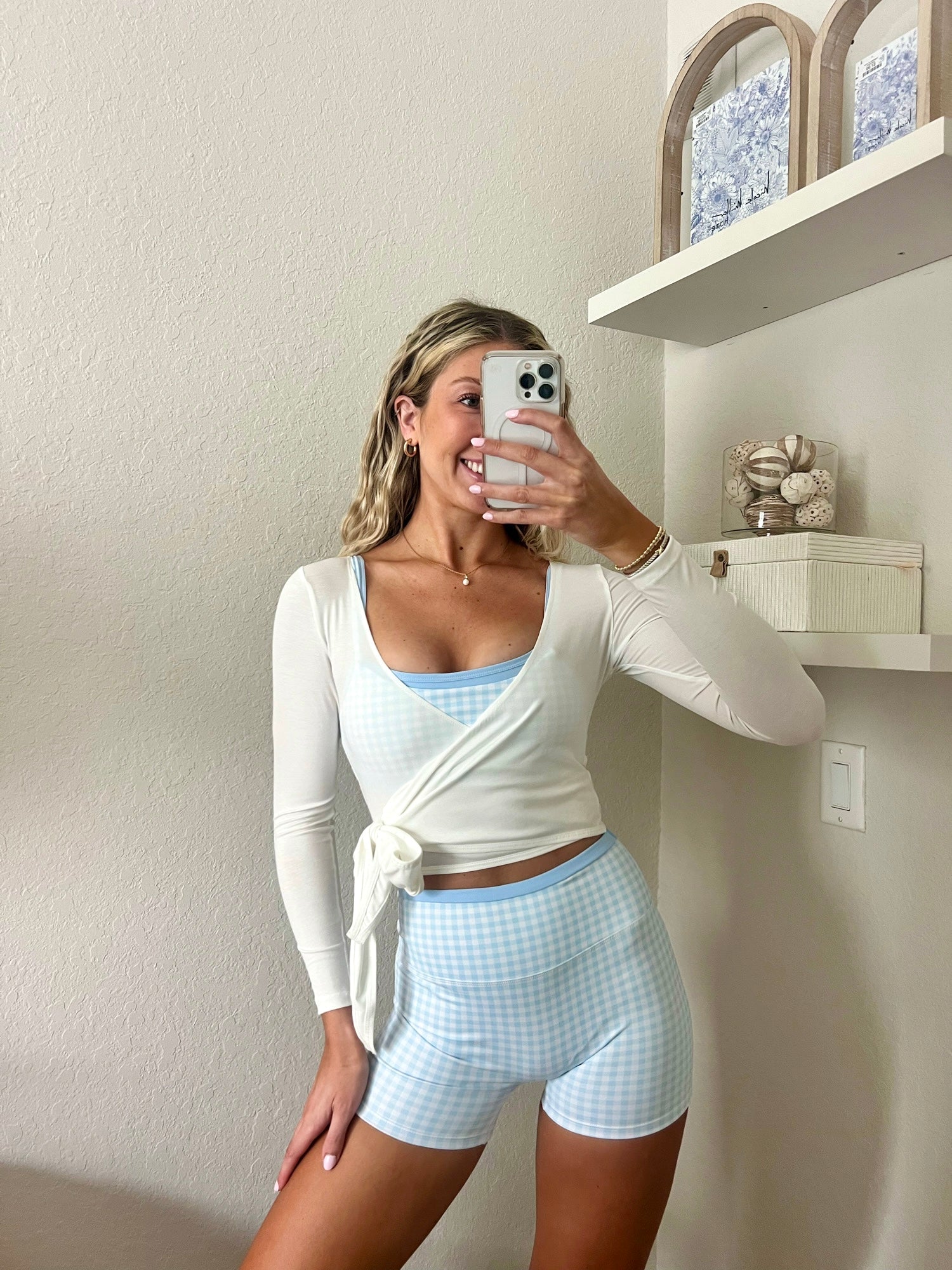 Woman taking a mirror selfie wearing a light blue and white gingham workout set, consisting of a sports bra and biker shorts, with a white long sleeve wrap top, in a home setting.