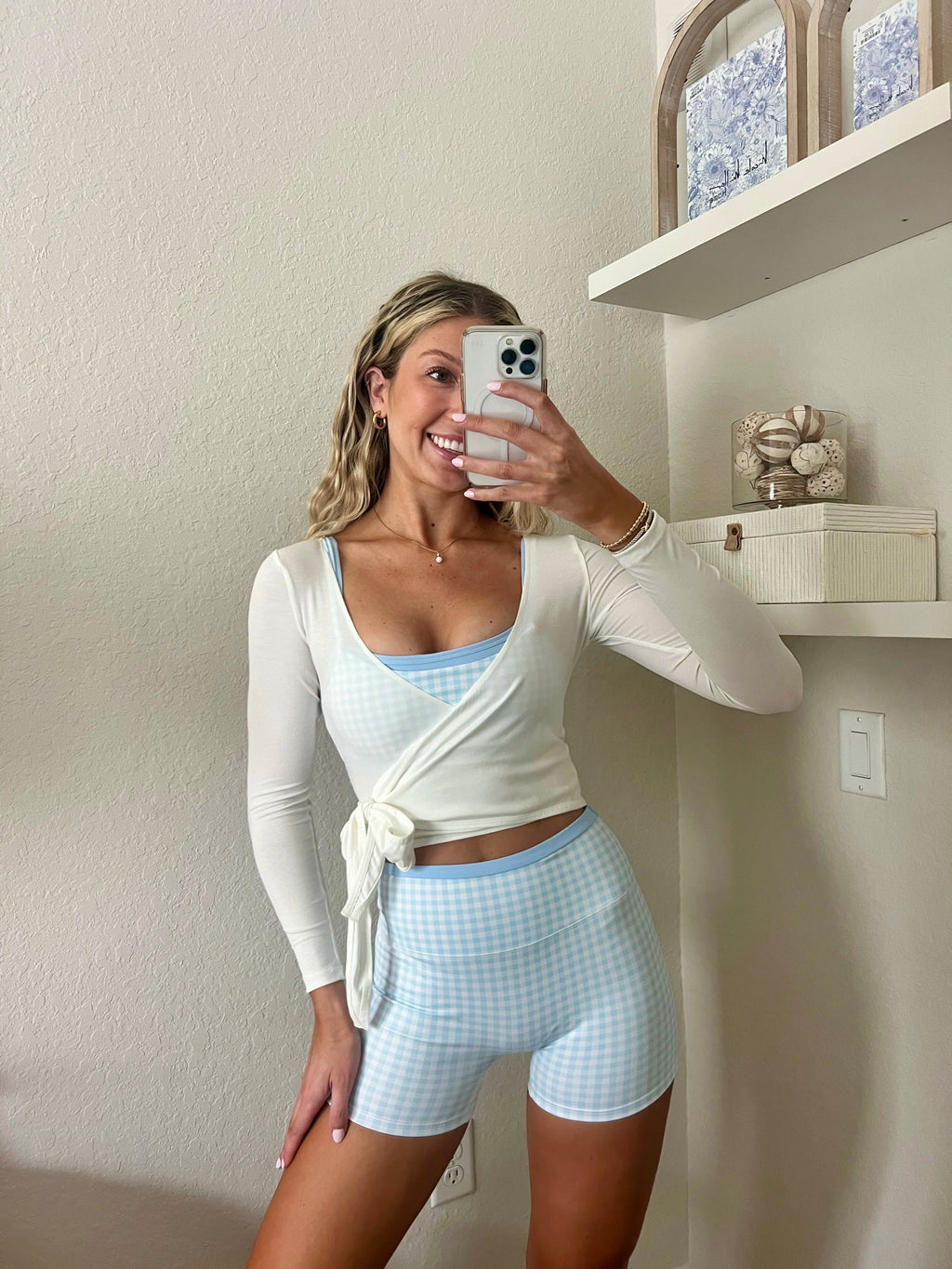 Woman taking a mirror selfie wearing a light blue and white gingham workout set, consisting of a sports bra and biker shorts, with a white long sleeve wrap top, in a home setting.