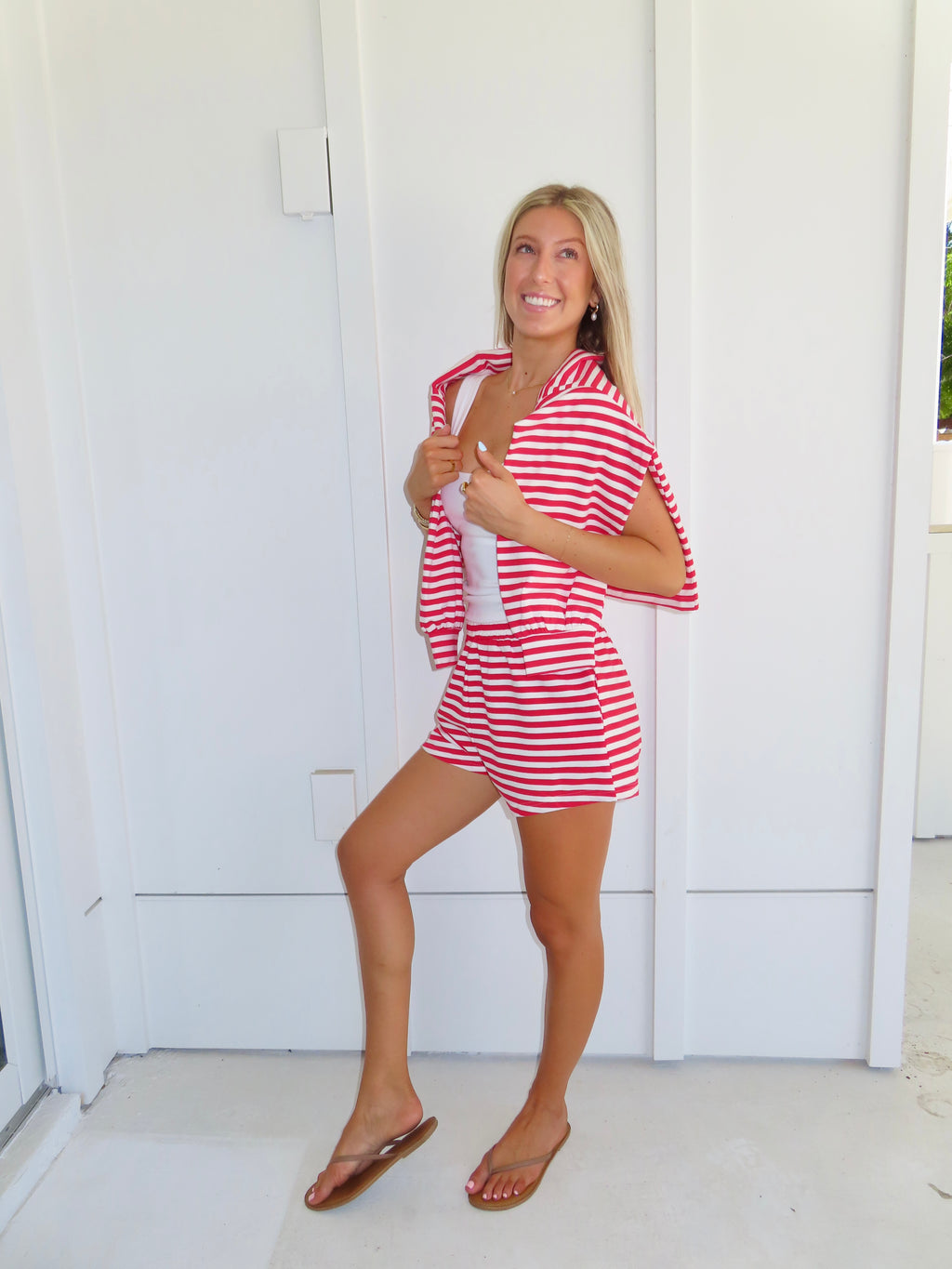 Woman wearing a red and white striped set with a white tank under, wearing a sandals against a white house.