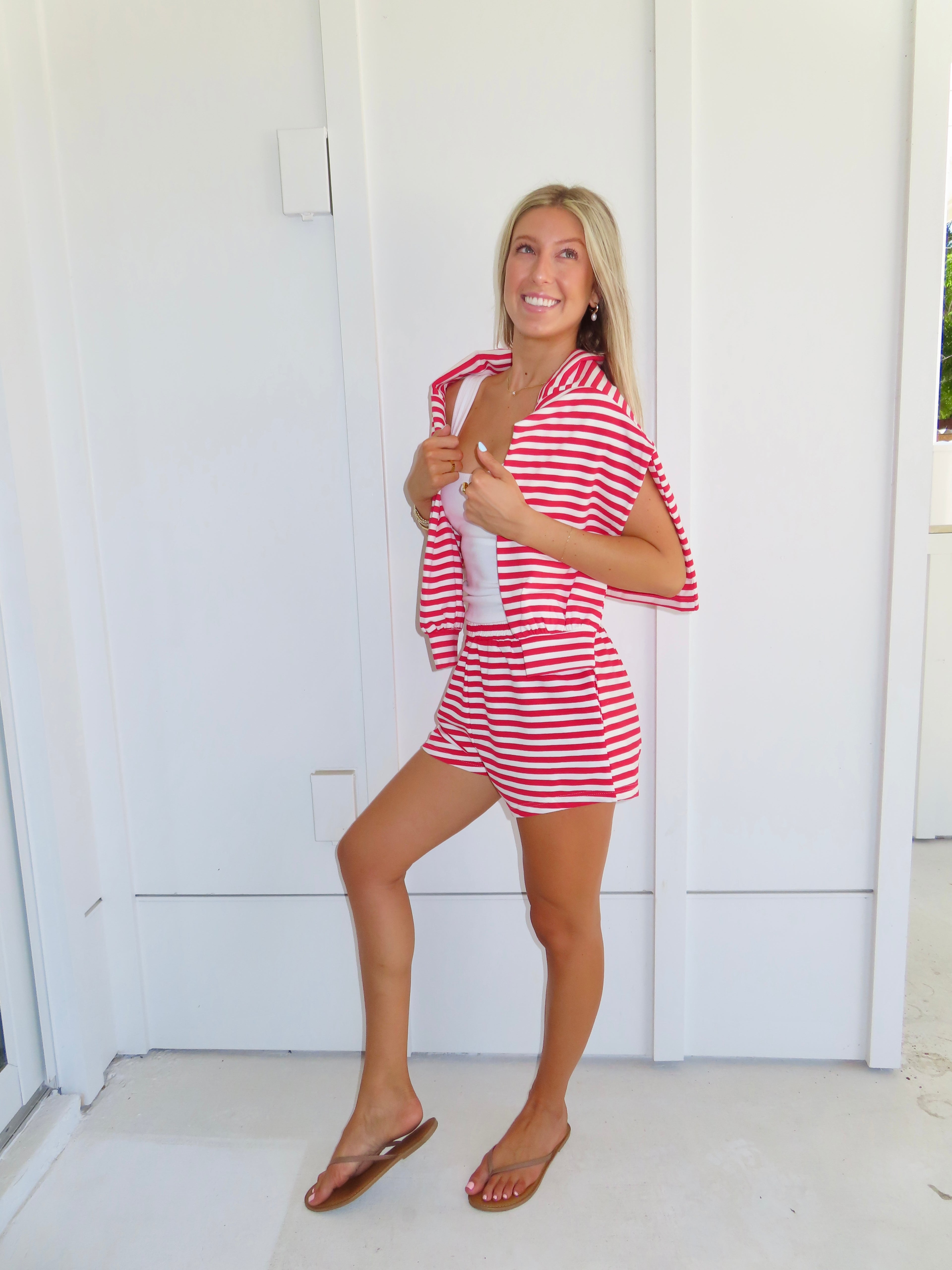 Woman wearing a red and white striped set with a white tank under, wearing a sandals against a white house.