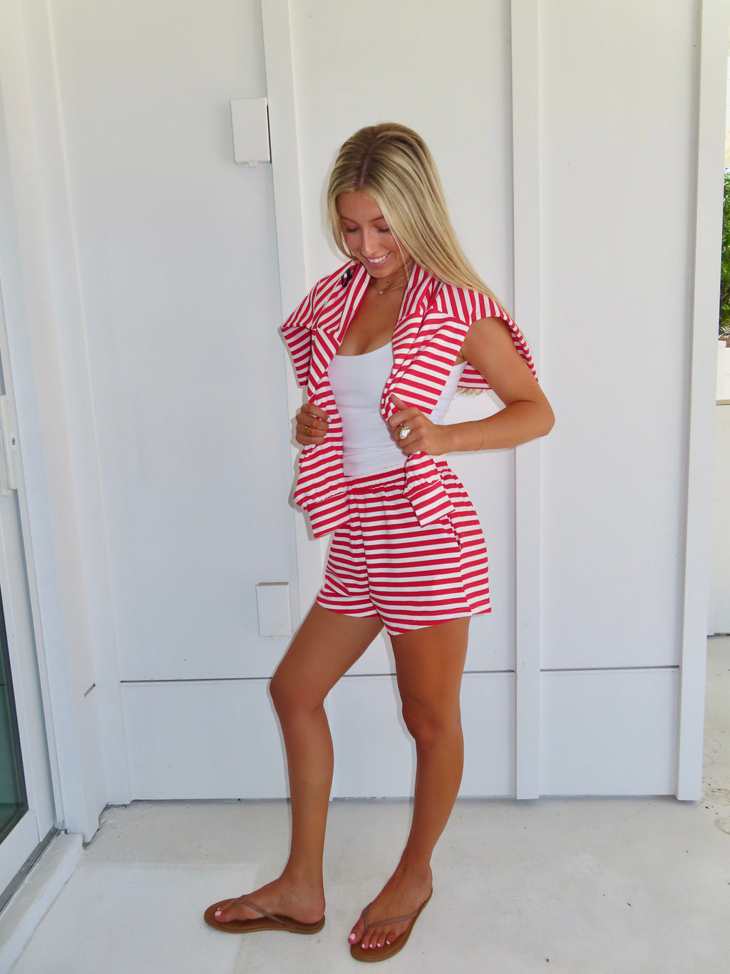Woman wearing a red and white striped set with a white tank under, wearing a sandals against a white house.