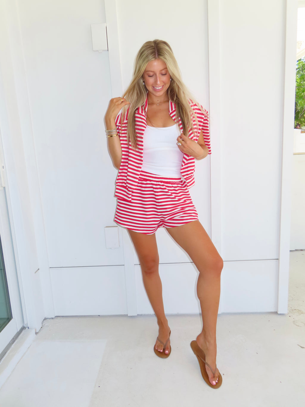 Woman wearing a red and white striped set with a white tank under, wearing a sandals against a white house.