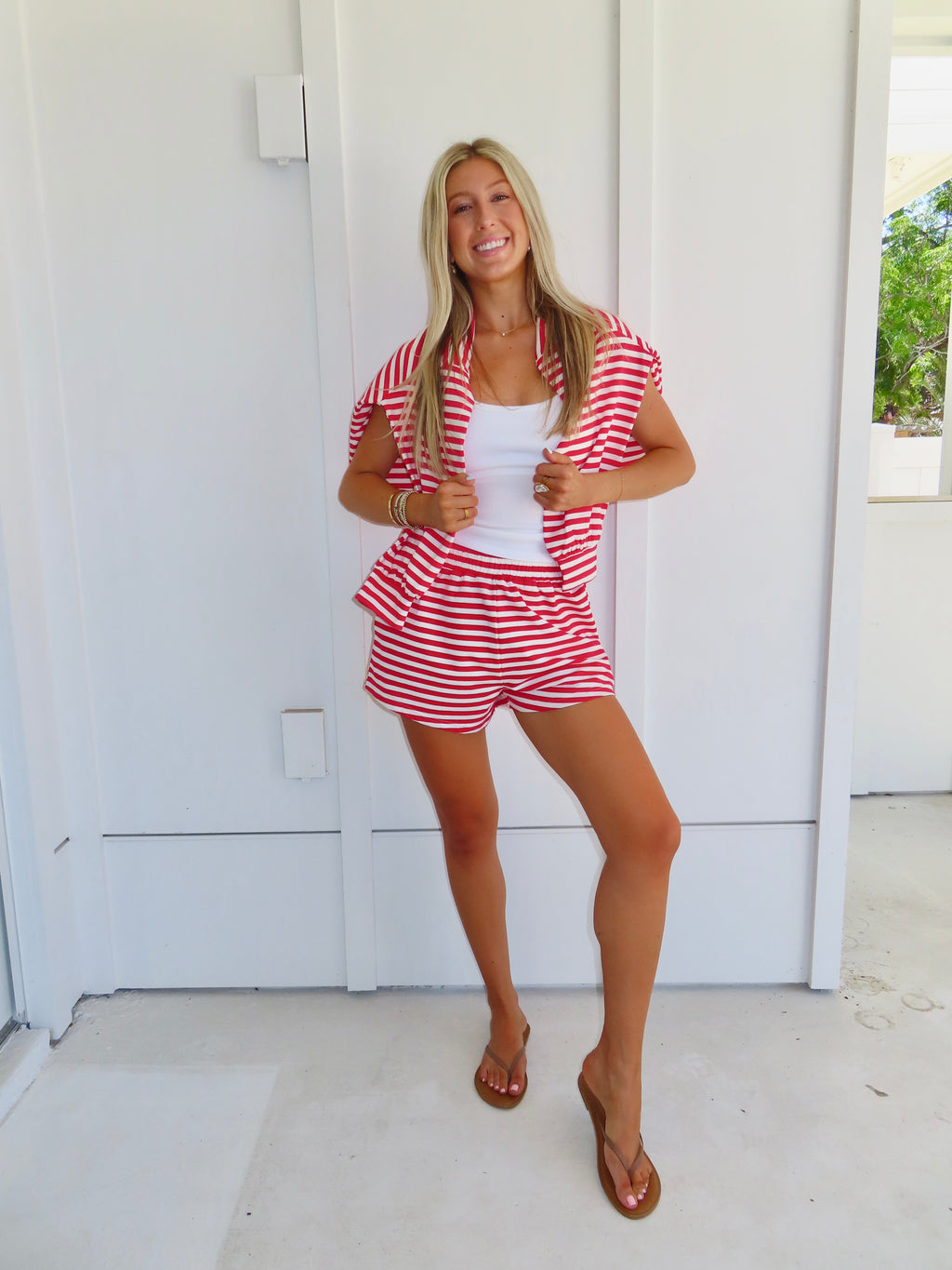 Woman wearing a red and white striped set with a white tank under, wearing a sandals against a white house.