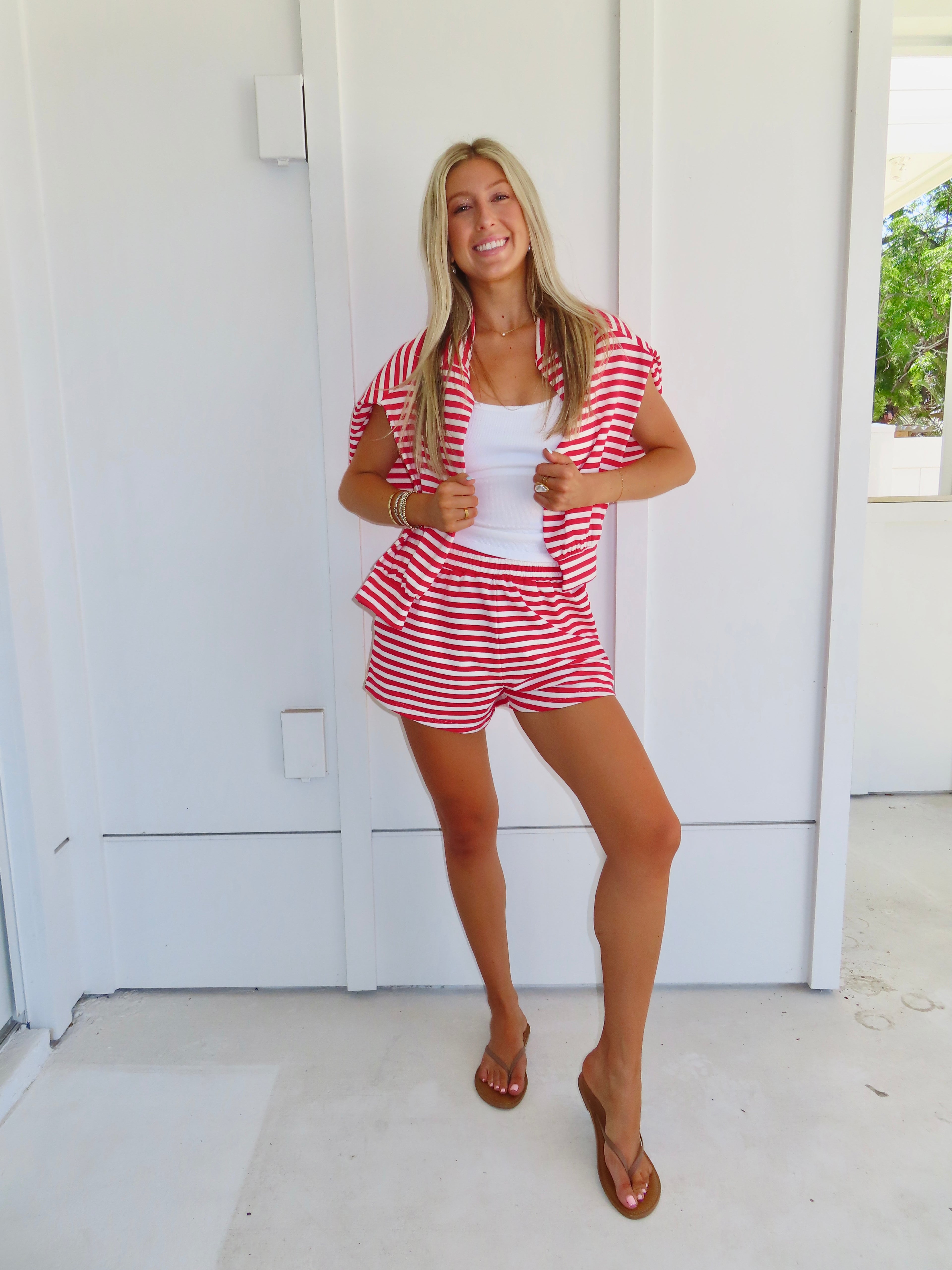 Woman wearing a red and white striped set with a white tank under, wearing a sandals against a white house.