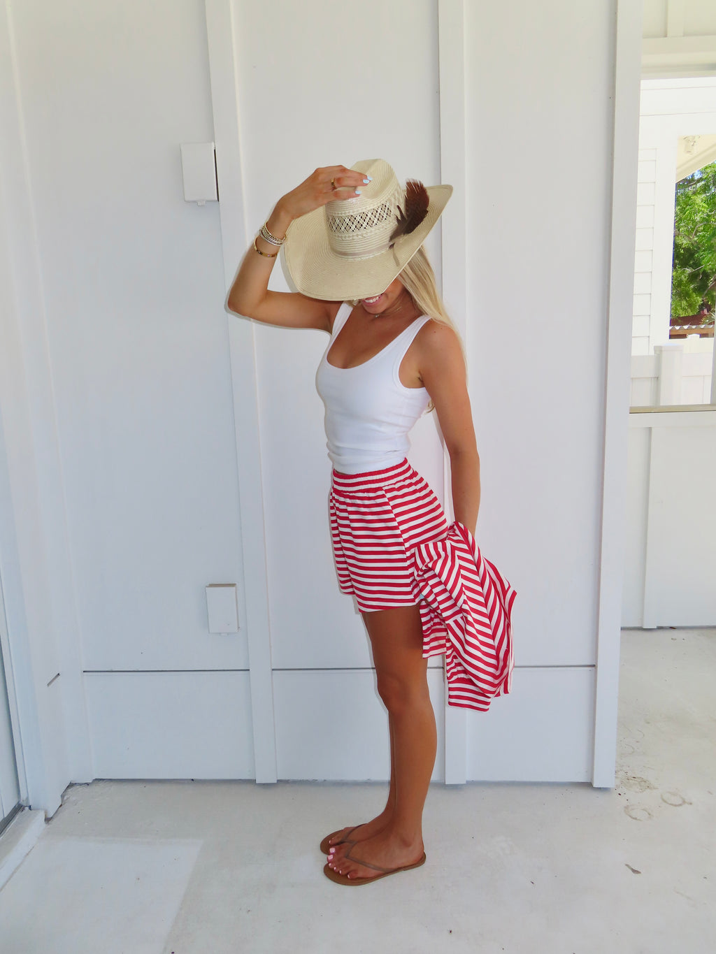 Woman wearing a red and white striped set with a white tank under, wearing a sandals and a cowgirl hat against a white house.