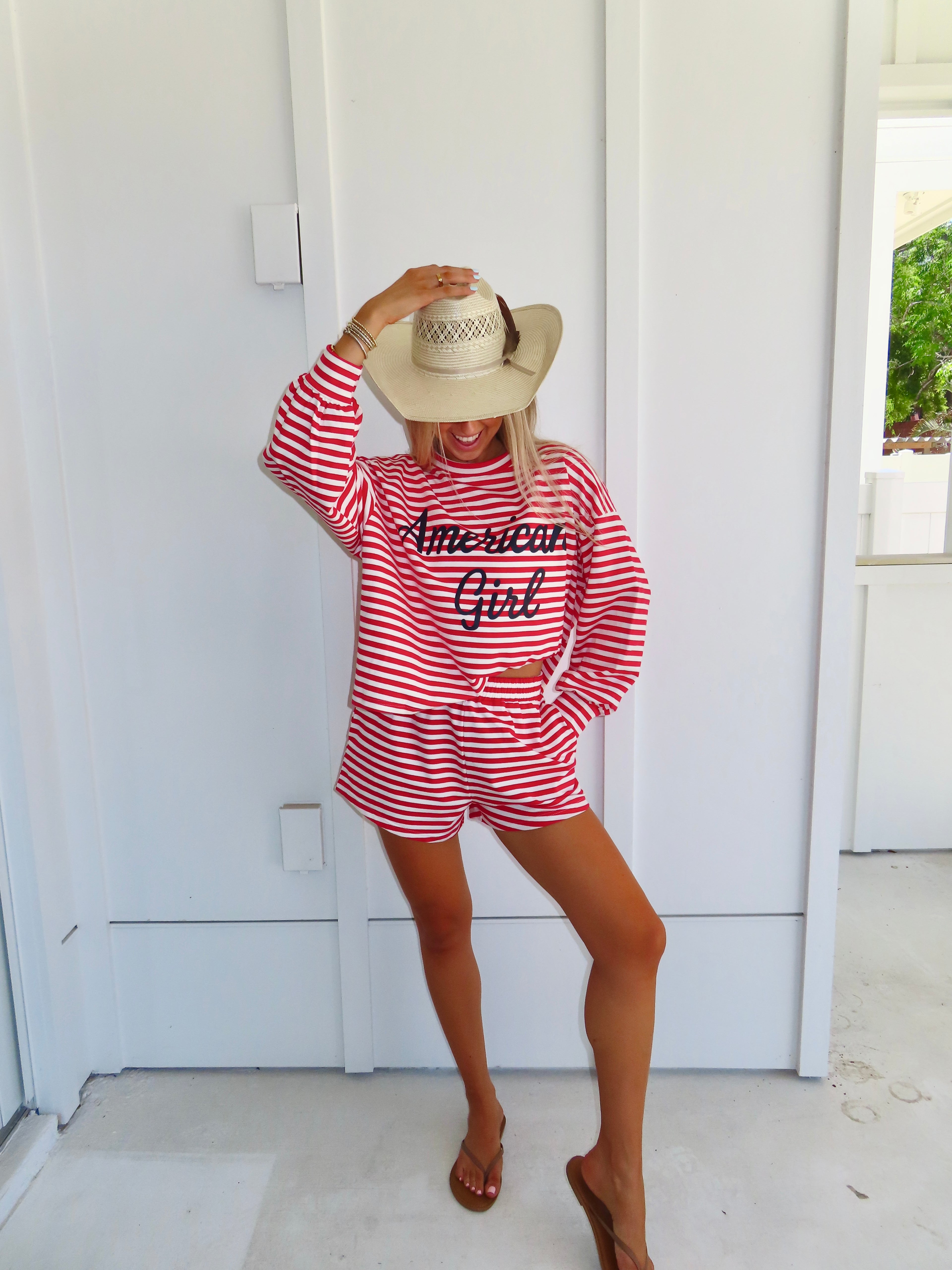 Woman wearing a red and white striped set that says "American girl" in navy, wearing a cowgirl hat and sandals.