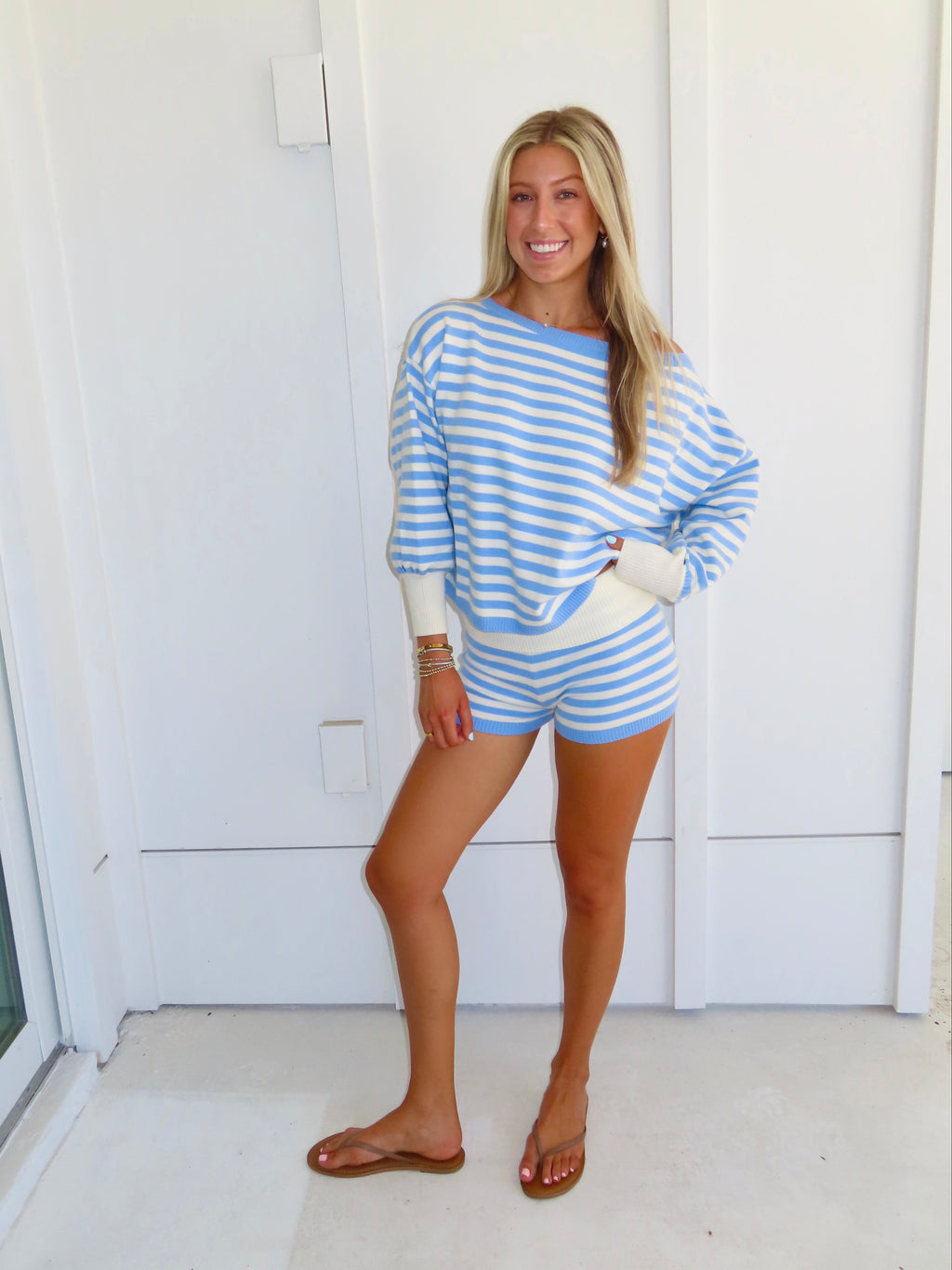 Woman standing against a white house wearing a blue and white striped shorts and sweater set, with sandals.