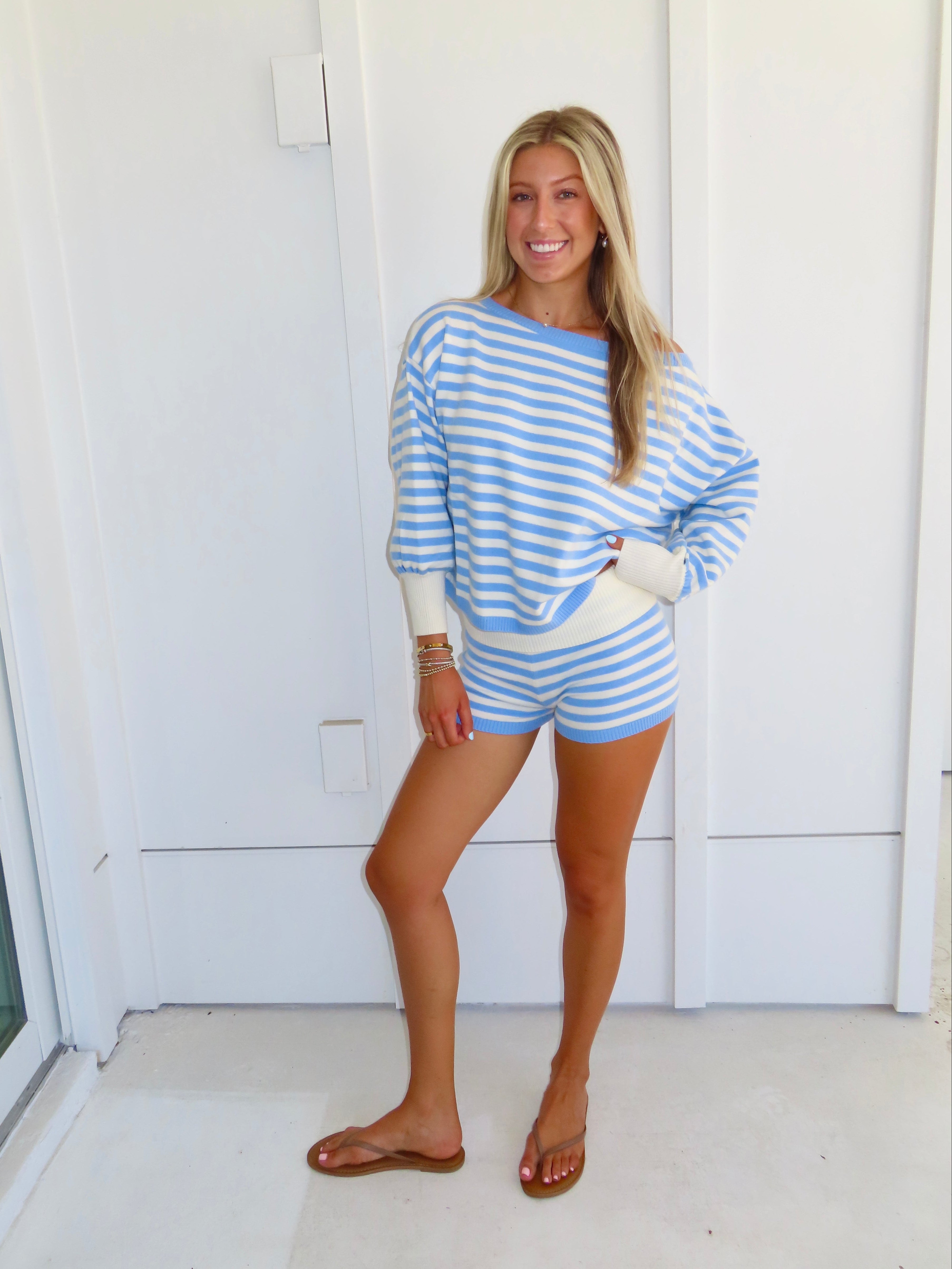 Woman standing against a white house wearing a blue and white striped shorts and sweater set, with sandals.