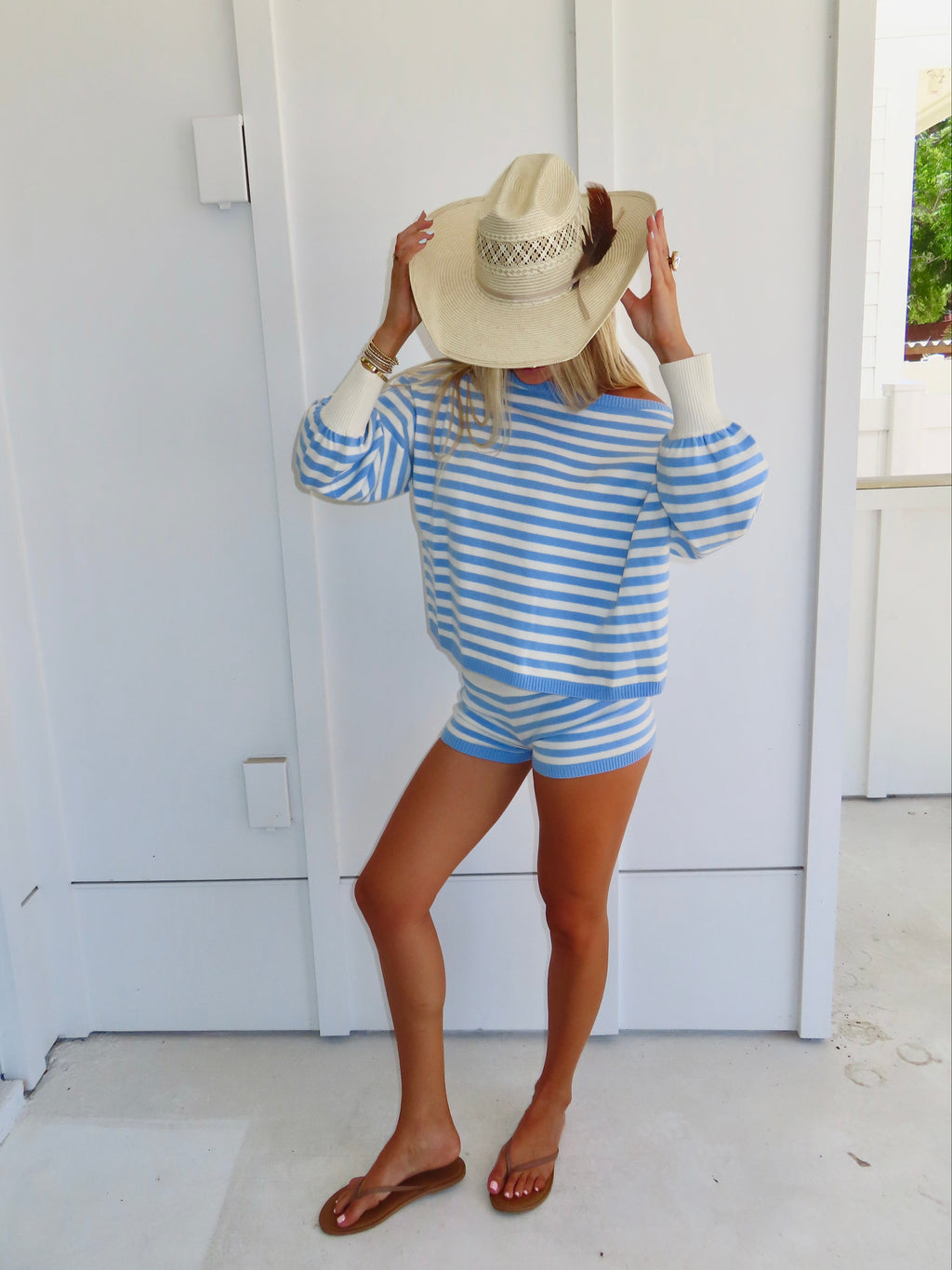 Woman standing against a white house wearing a blue and white striped shorts and sweater set, with sandals and a cowgirl hat.