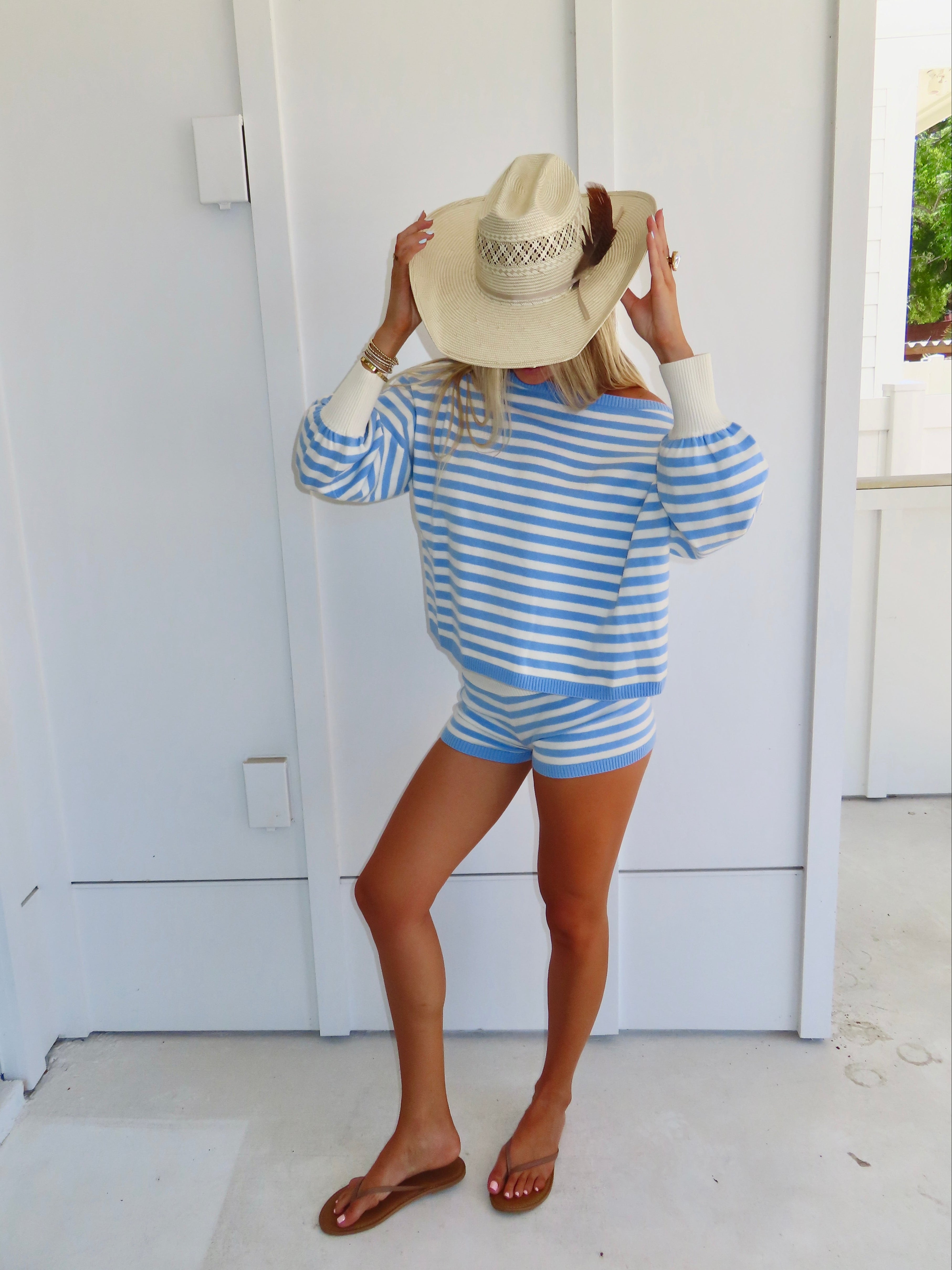 Woman standing against a white house wearing a blue and white striped shorts and sweater set, with sandals and a cowgirl hat.