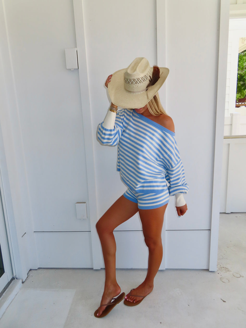 Woman standing against a white house wearing a blue and white striped shorts and sweater set, with sandals and a cowgirl hat.