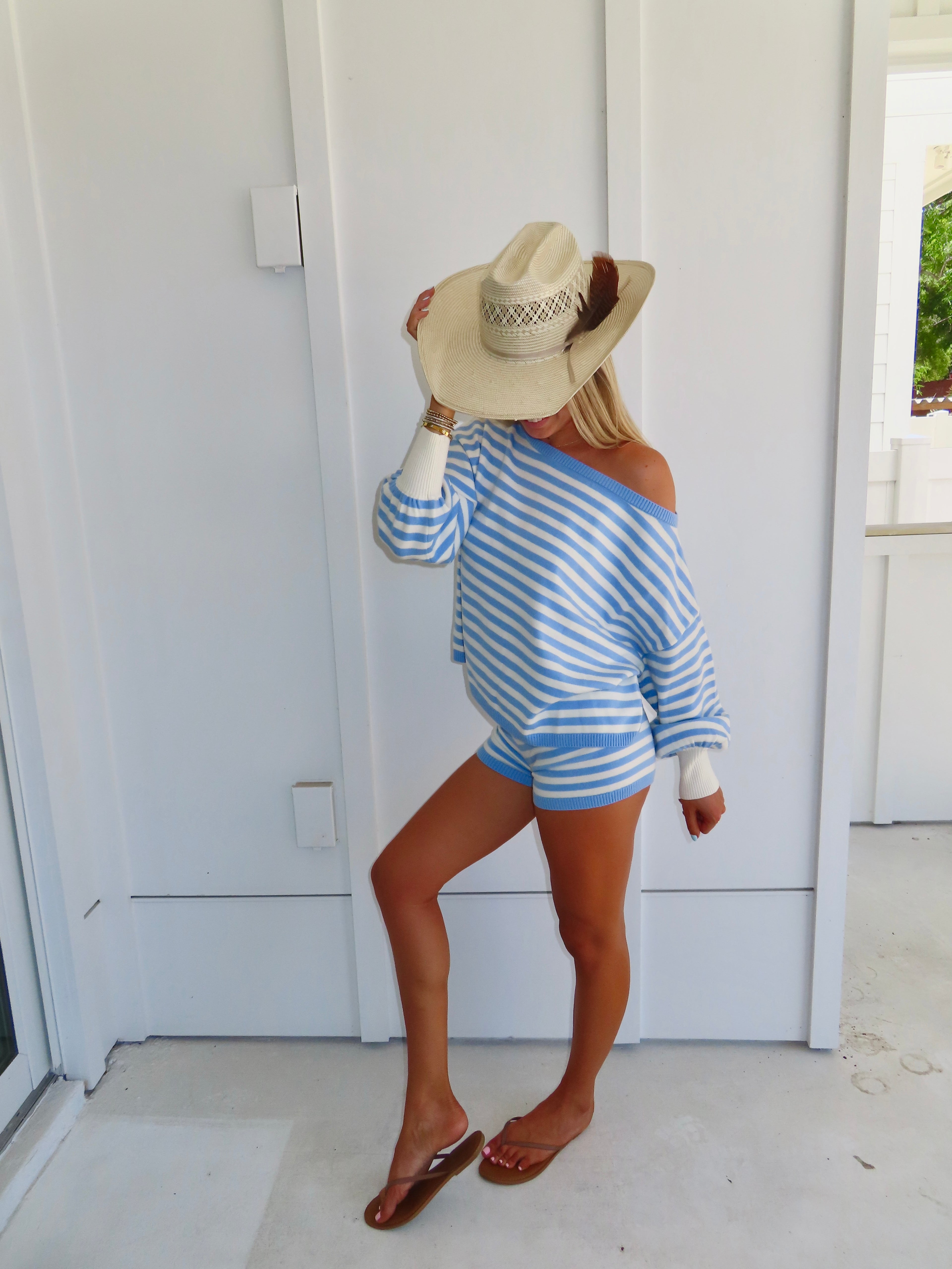 Woman standing against a white house wearing a blue and white striped shorts and sweater set, with sandals and a cowgirl hat.