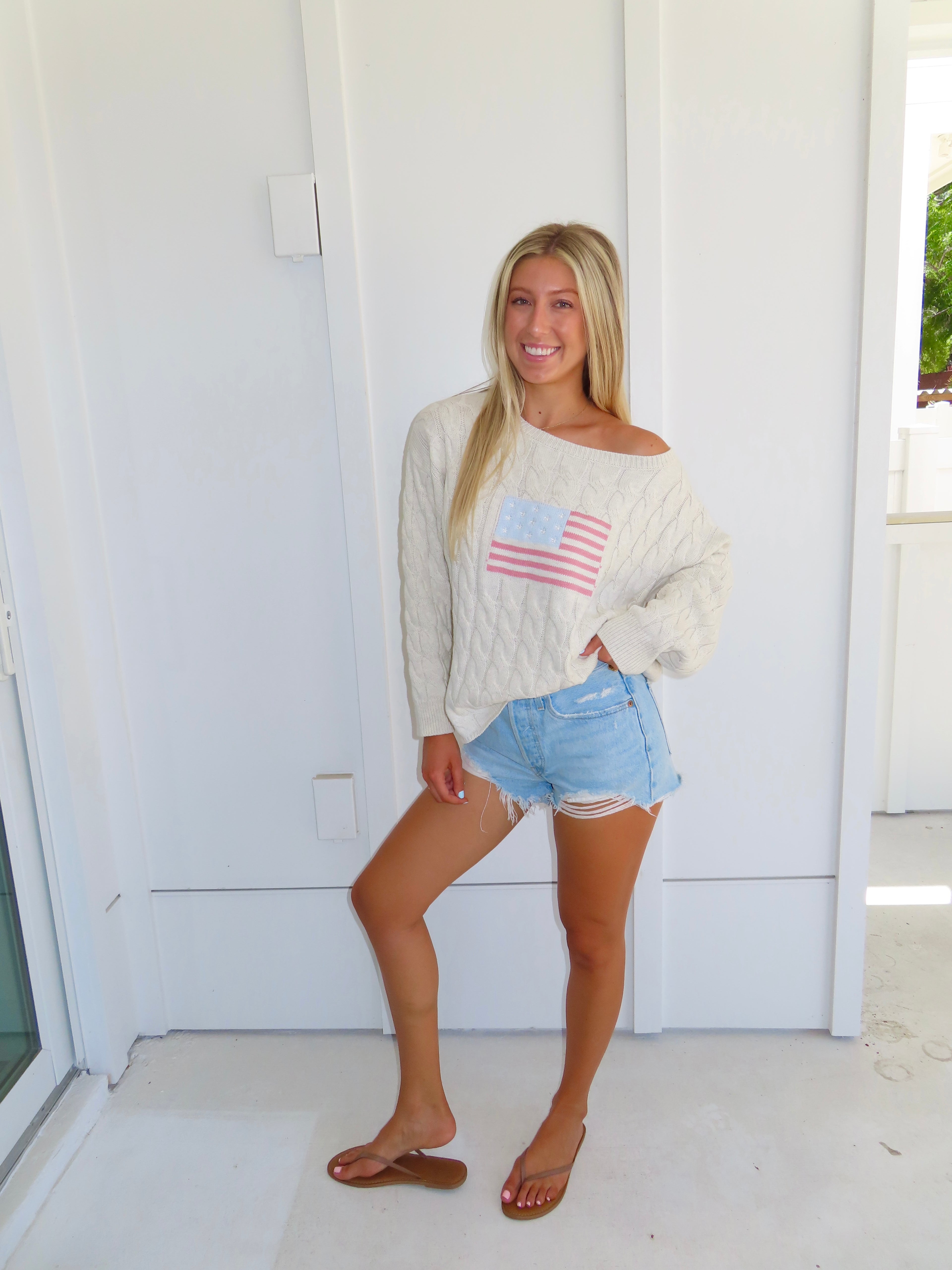 Woman wearing an oatmeal American flag sweater with denim shorts and sandals against a white house.