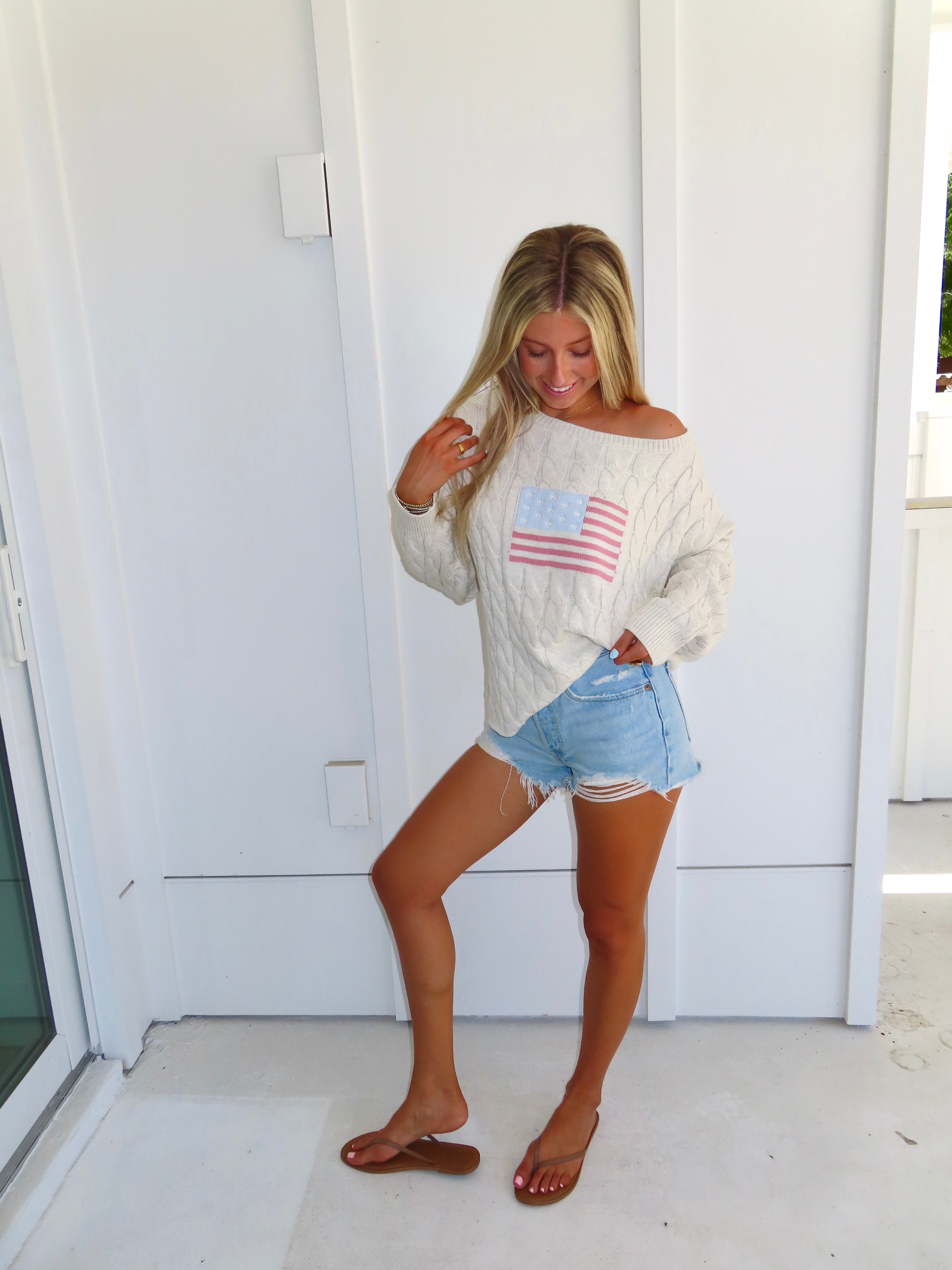 Woman wearing an oatmeal American flag sweater with denim shorts and sandals against a white house.