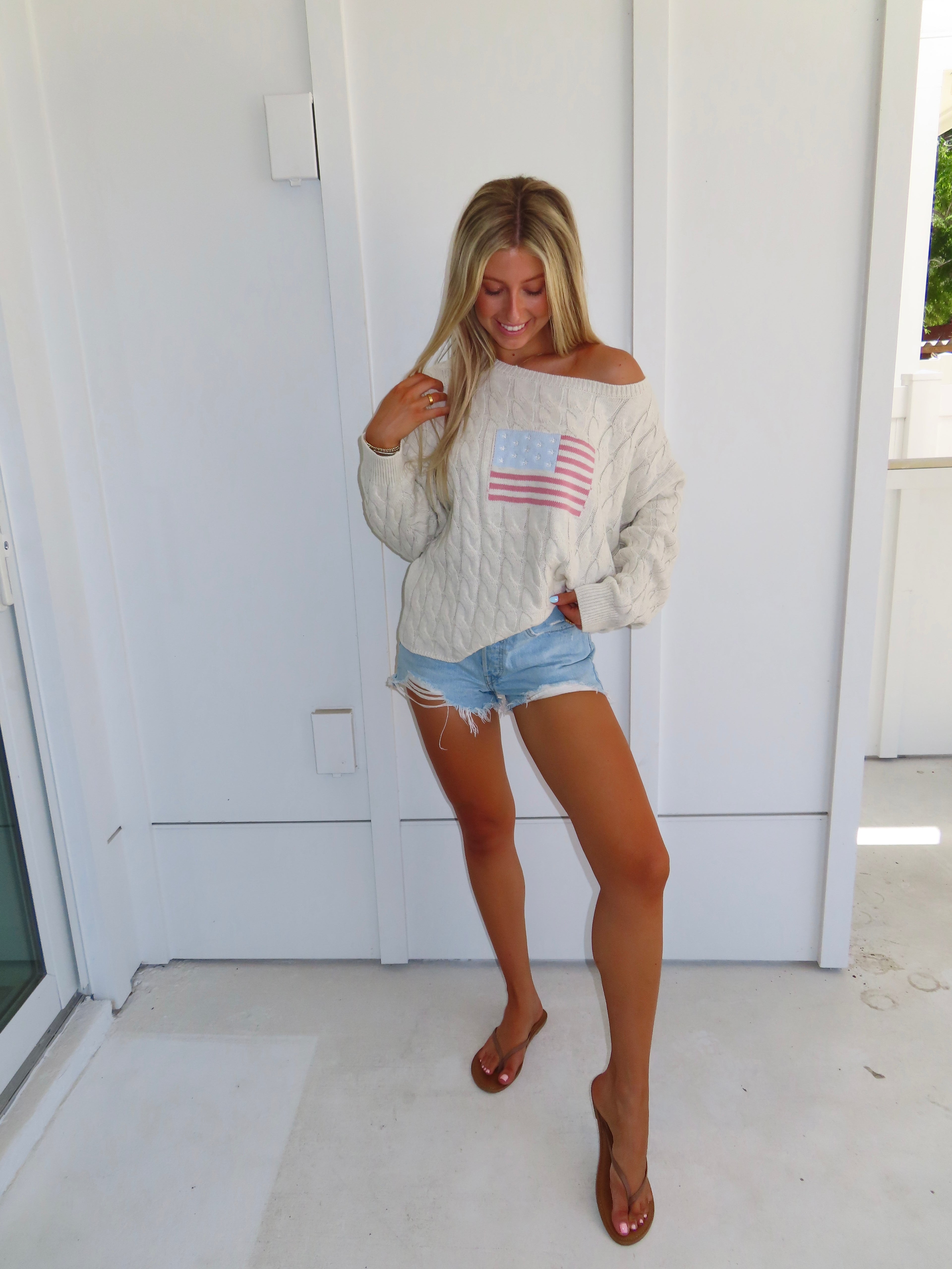 Woman wearing an oatmeal American flag sweater with denim shorts and sandals against a white house.