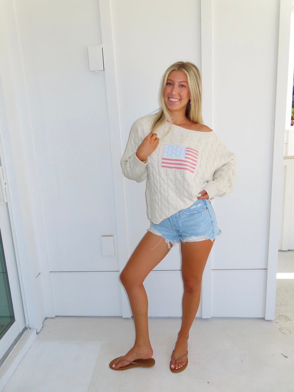 Woman wearing an oatmeal American flag sweater with denim shorts and sandals against a white house.