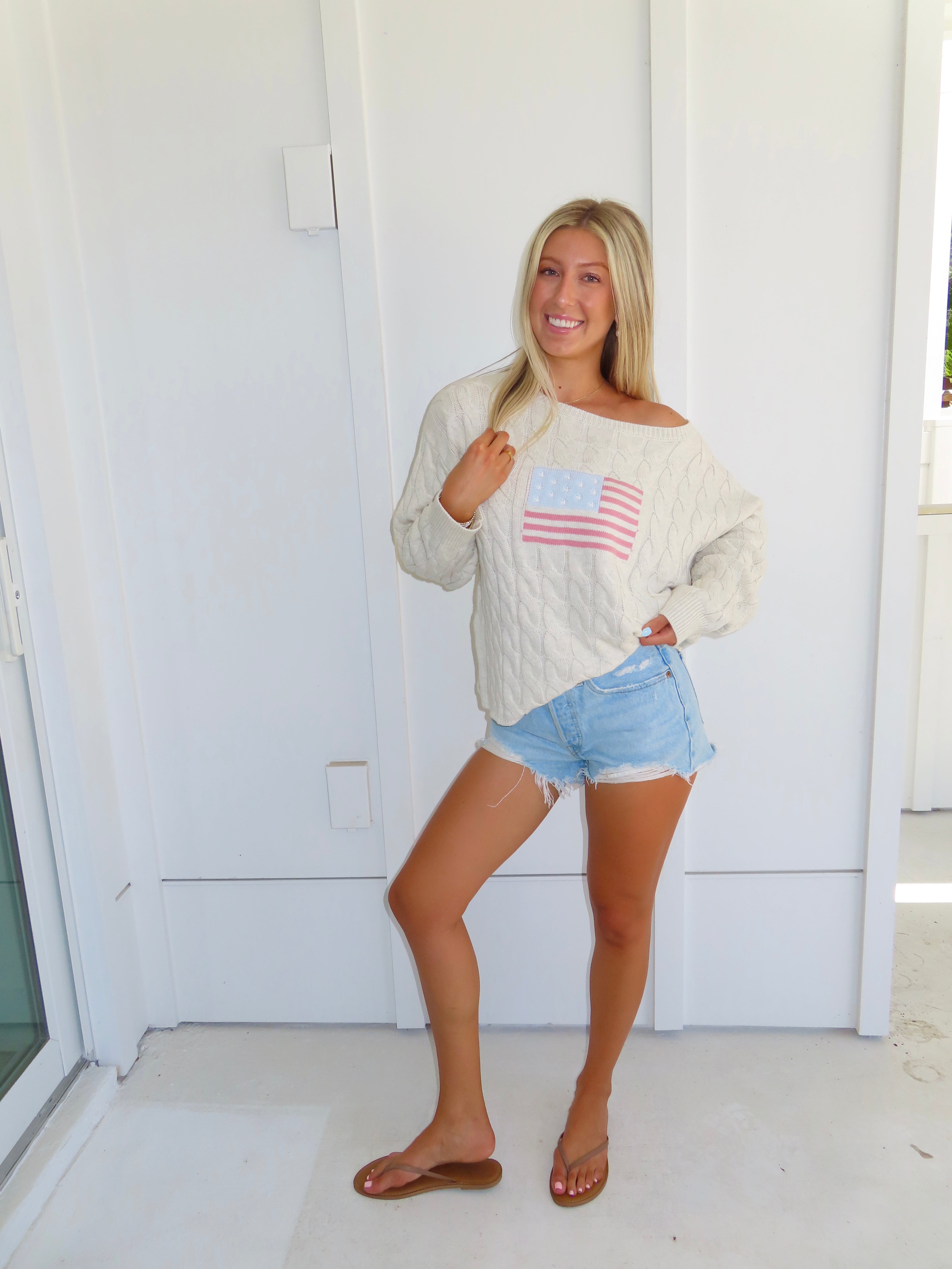 Woman wearing an oatmeal American flag sweater with denim shorts and sandals against a white house.