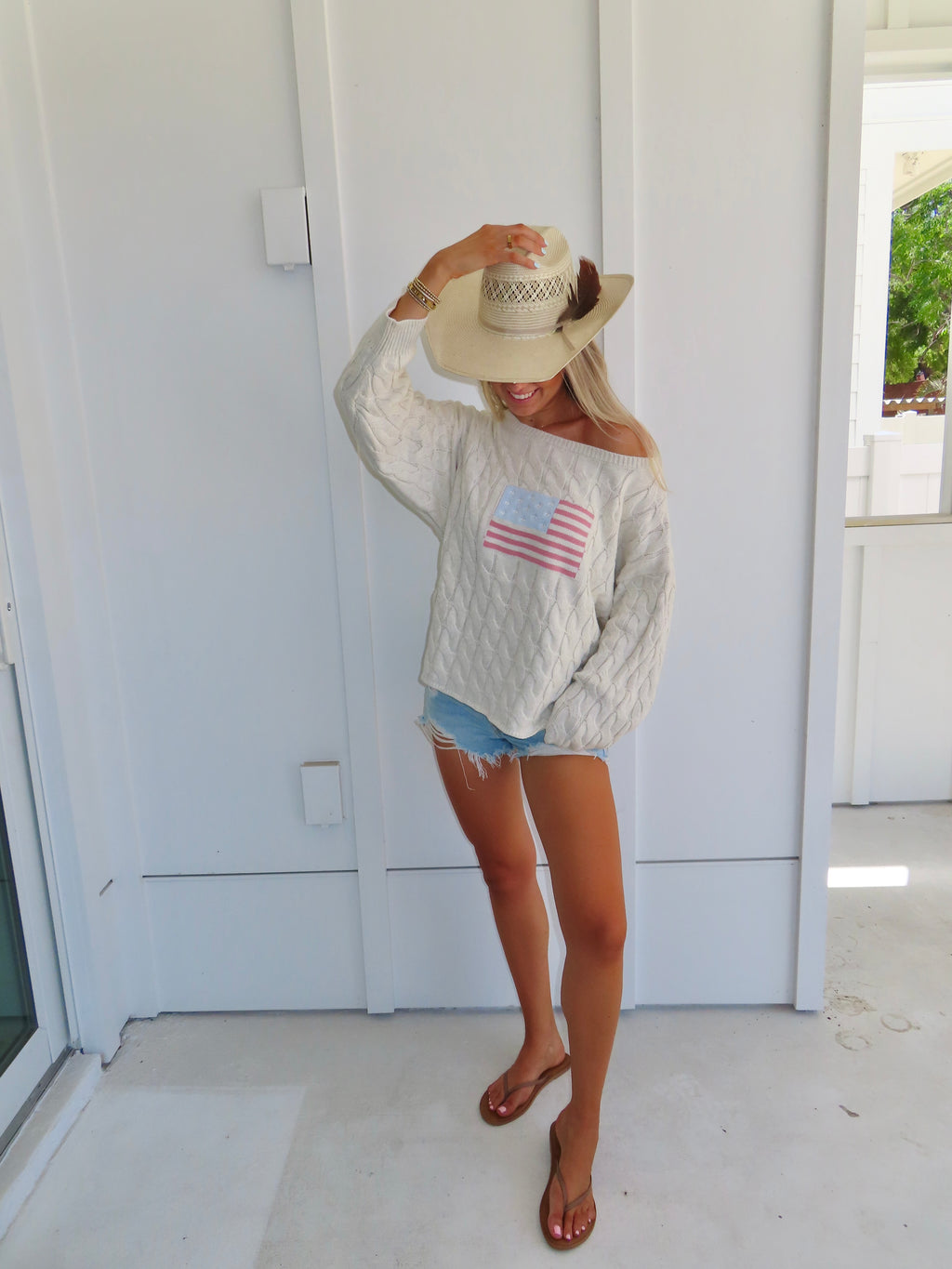 Woman wearing an oatmeal American flag sweater with denim shorts and sandals with a hat against a white house.
