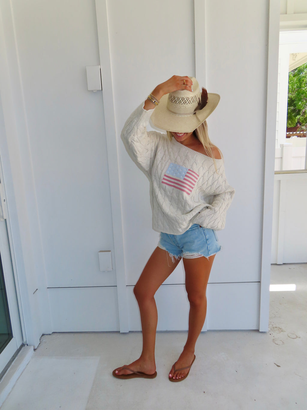 Woman wearing an oatmeal American flag sweater with denim shorts and sandals with a cowgirl hat against a white house.