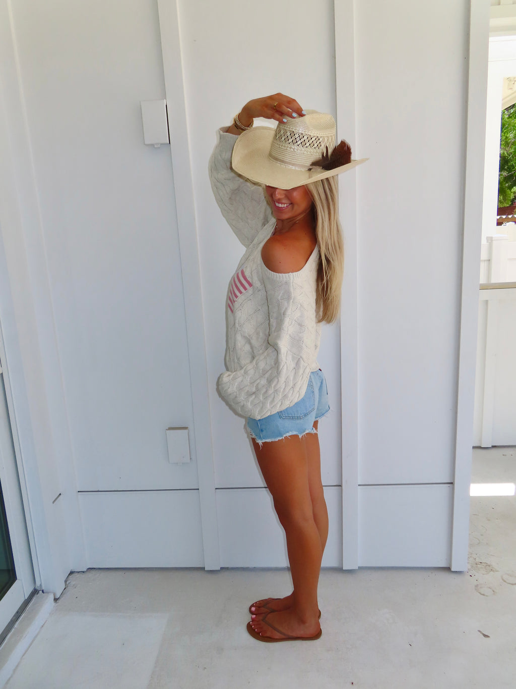 Woman wearing an oatmeal American flag sweater with denim shorts and sandals with a cowgirl hat against a white house.