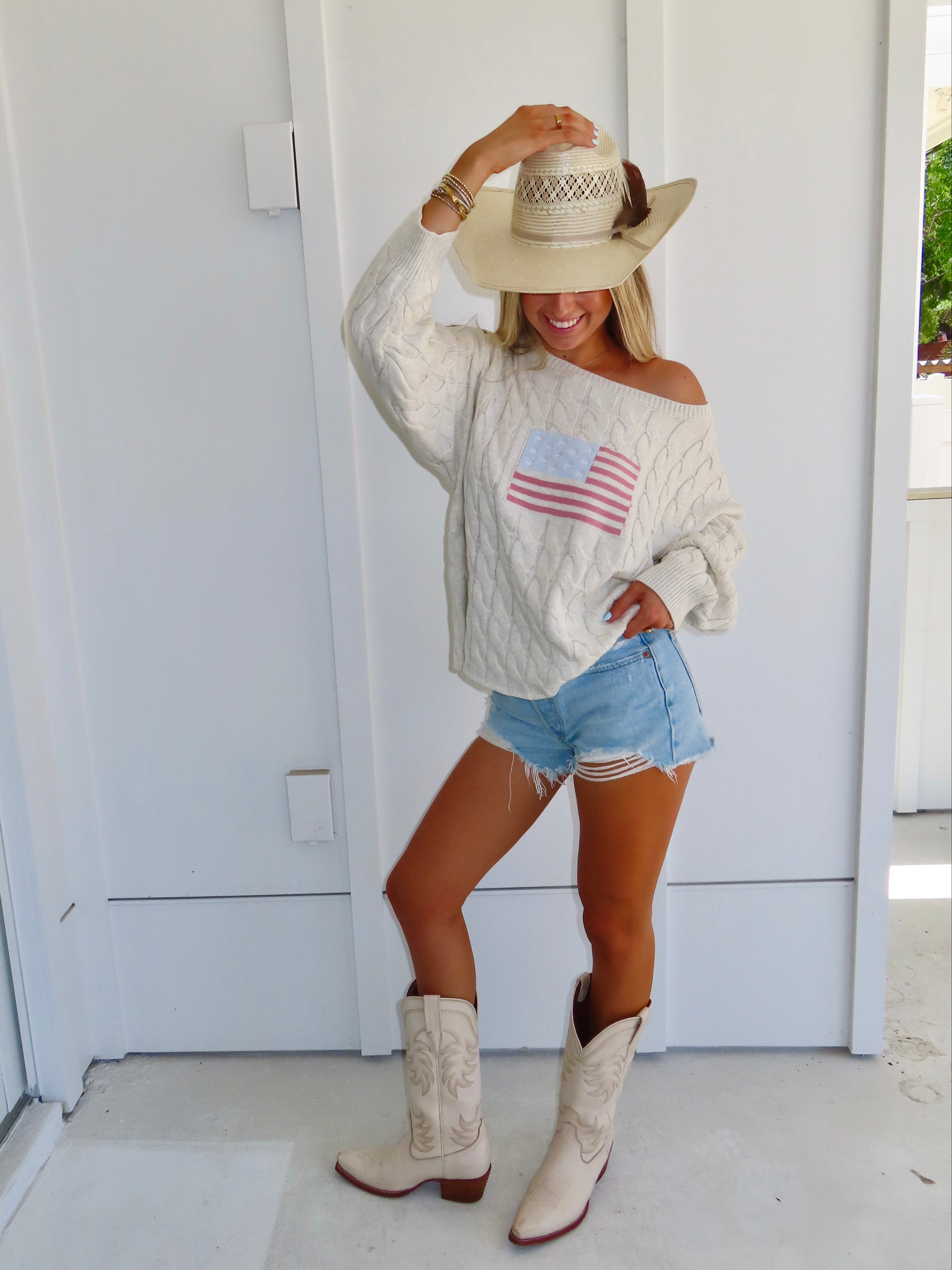Woman wearing an oatmeal American flag sweater with denim shorts and cowgirl boots with a hat against a white house.