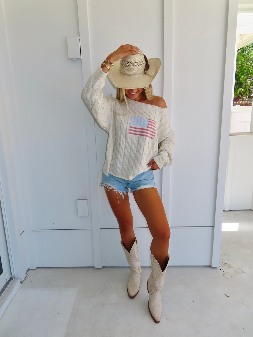 Woman wearing an oatmeal American flag sweater with denim shorts and cowgirl boots with a hat against a white house.