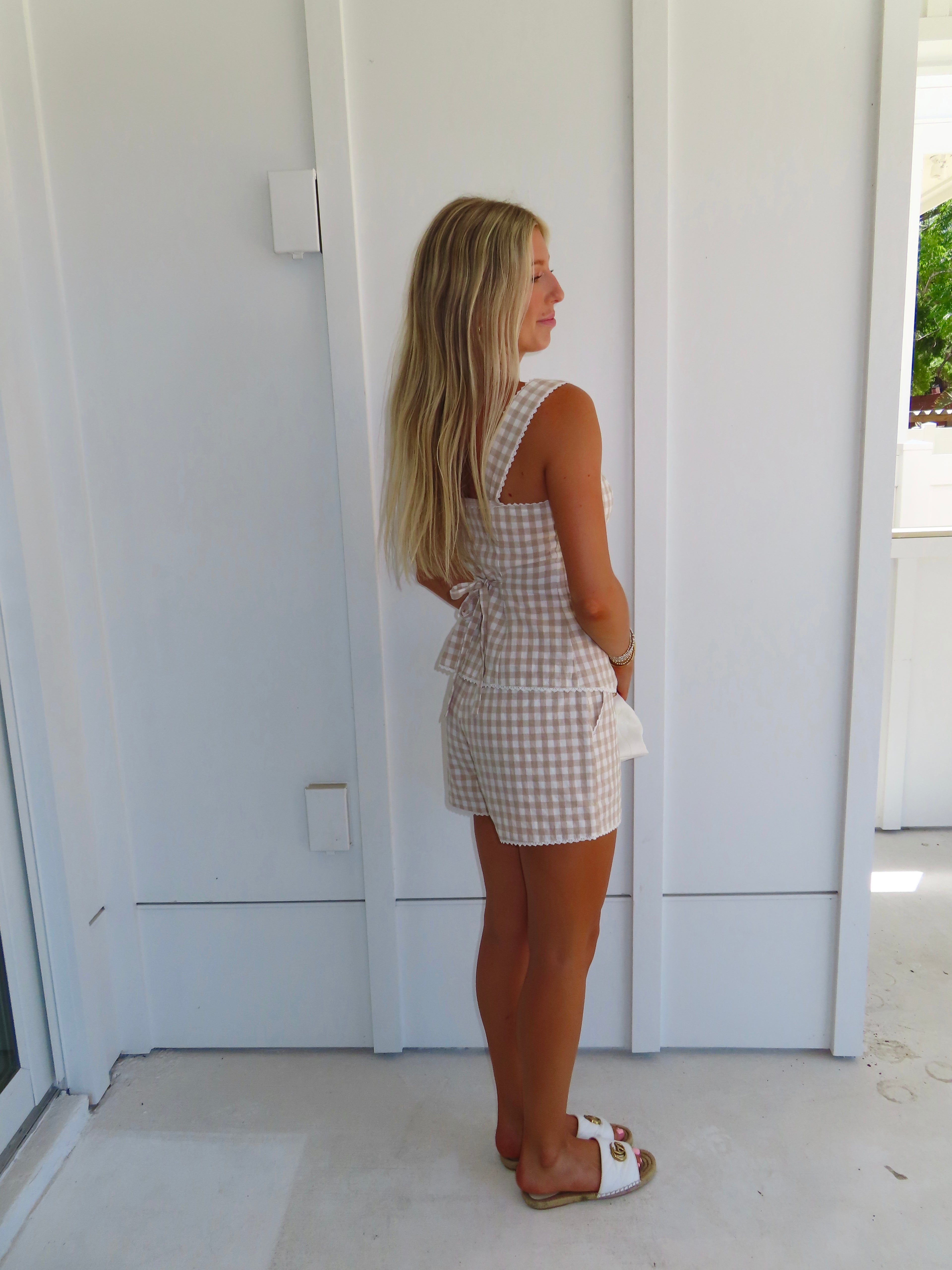 Woman wearing a tan and white gingham tank and shorts set with sandals and a purse, standing in front of a white house.