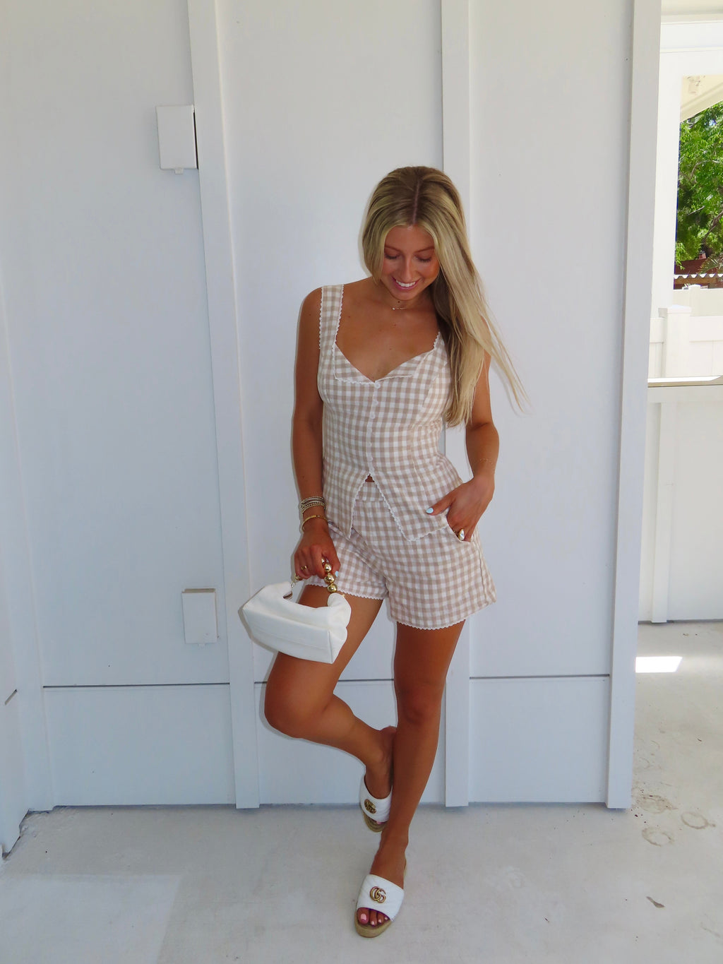 Woman wearing a tan and white gingham tank and shorts set with sandals and a purse, standing in front of a white house.