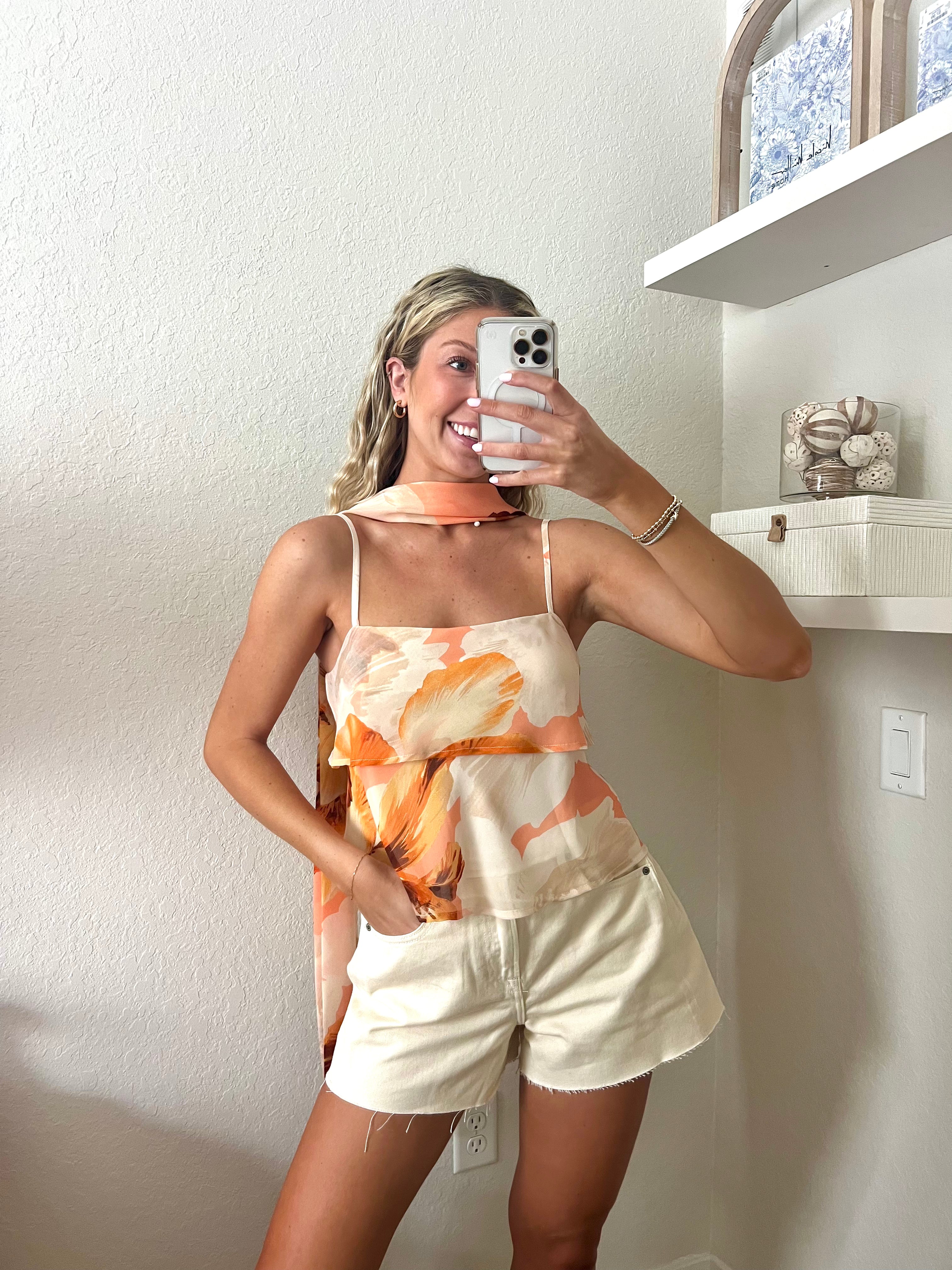 Woman taking a mirror selfie wearing a tank top with orange floral pattern, a neck scarf, and cream shorts.