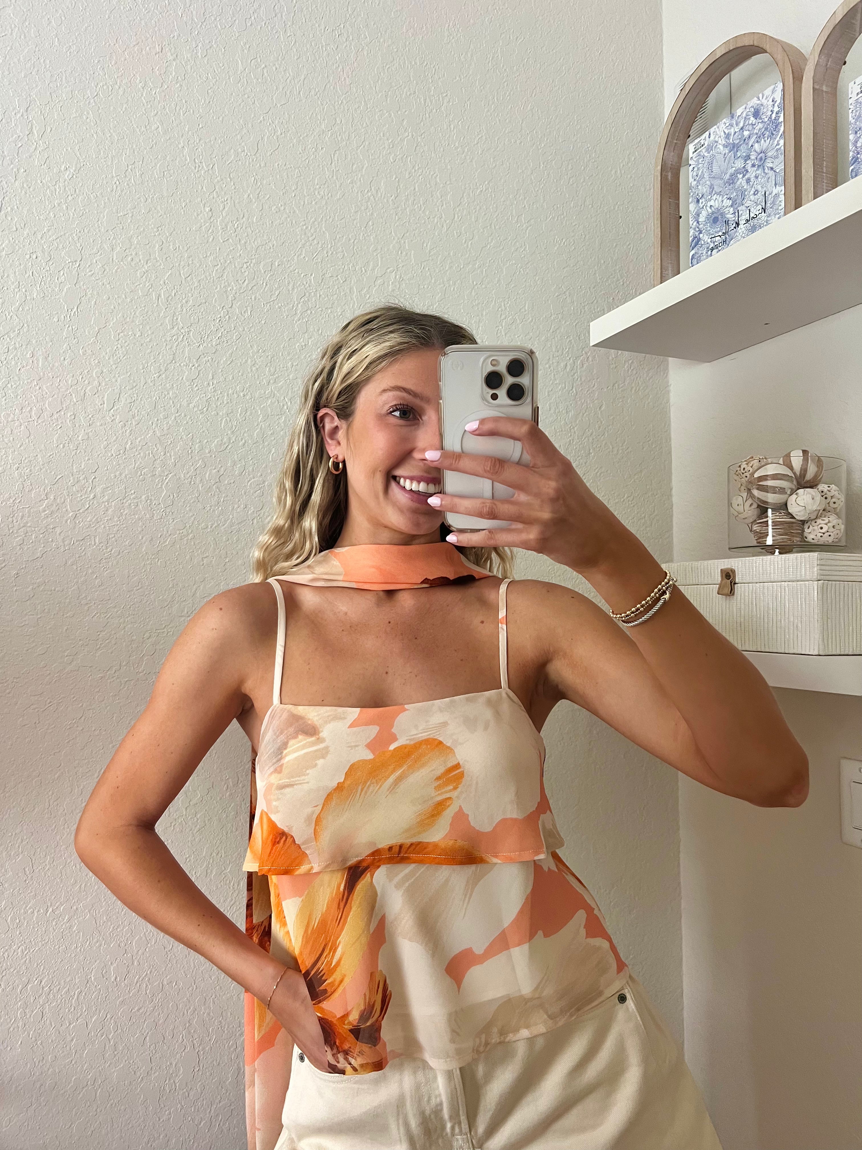 Woman taking a mirror selfie wearing a tank top with orange floral pattern, a neck scarf, and cream shorts.