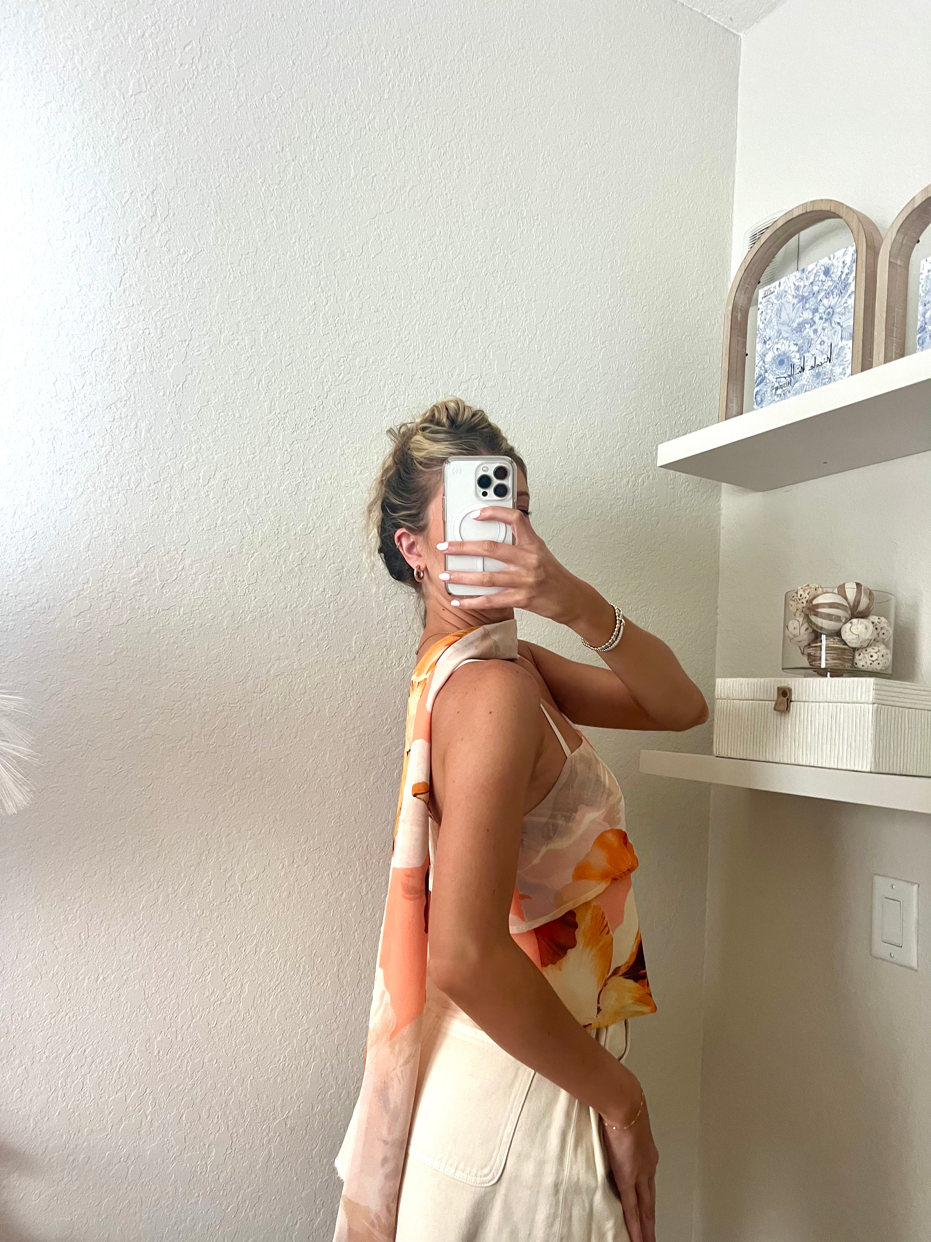 Woman taking a mirror selfie wearing a tank top with orange floral pattern, a neck scarf, and cream shorts.