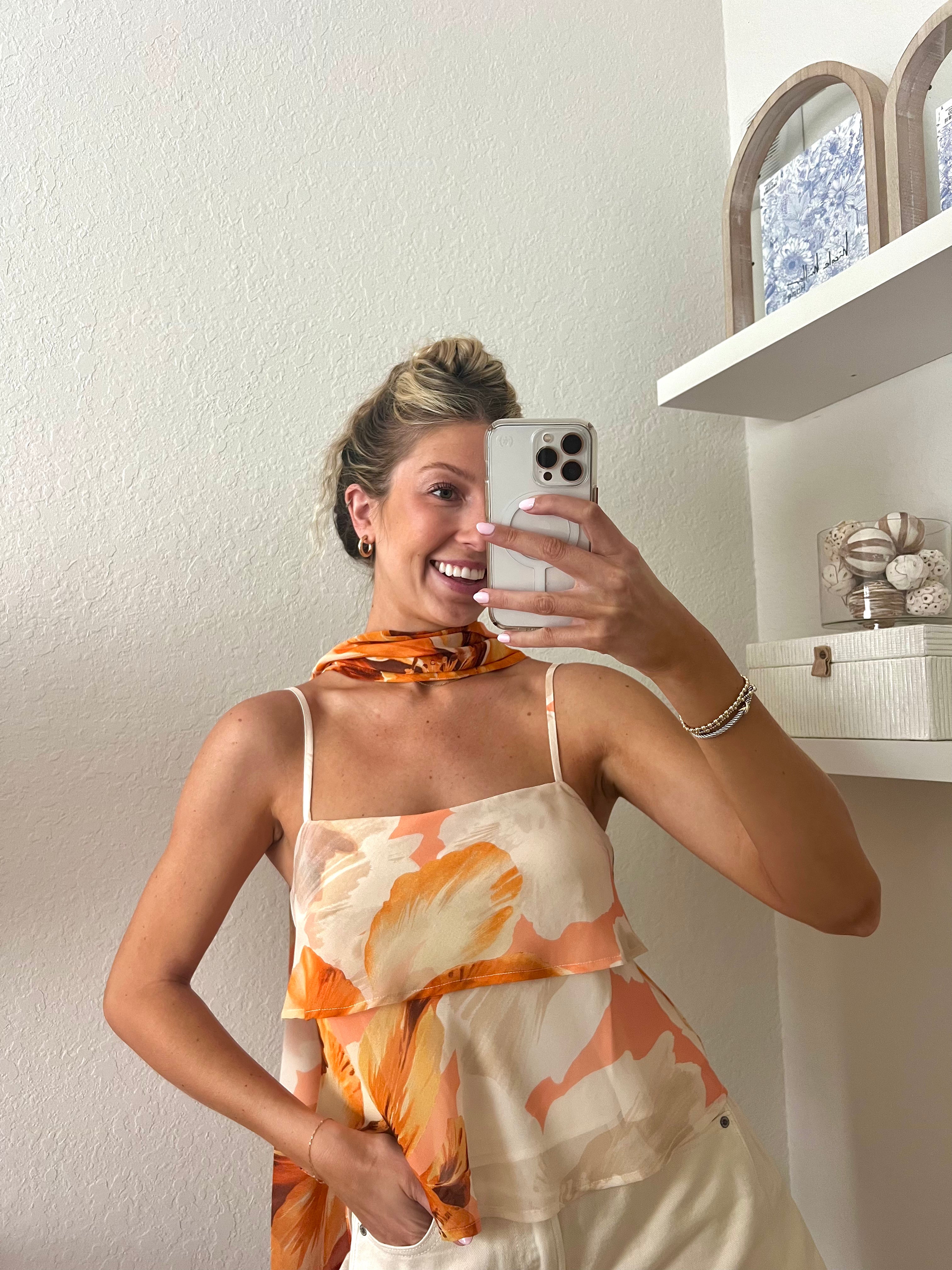 Woman taking a mirror selfie wearing a tank top with orange floral pattern, a neck scarf, and cream shorts.