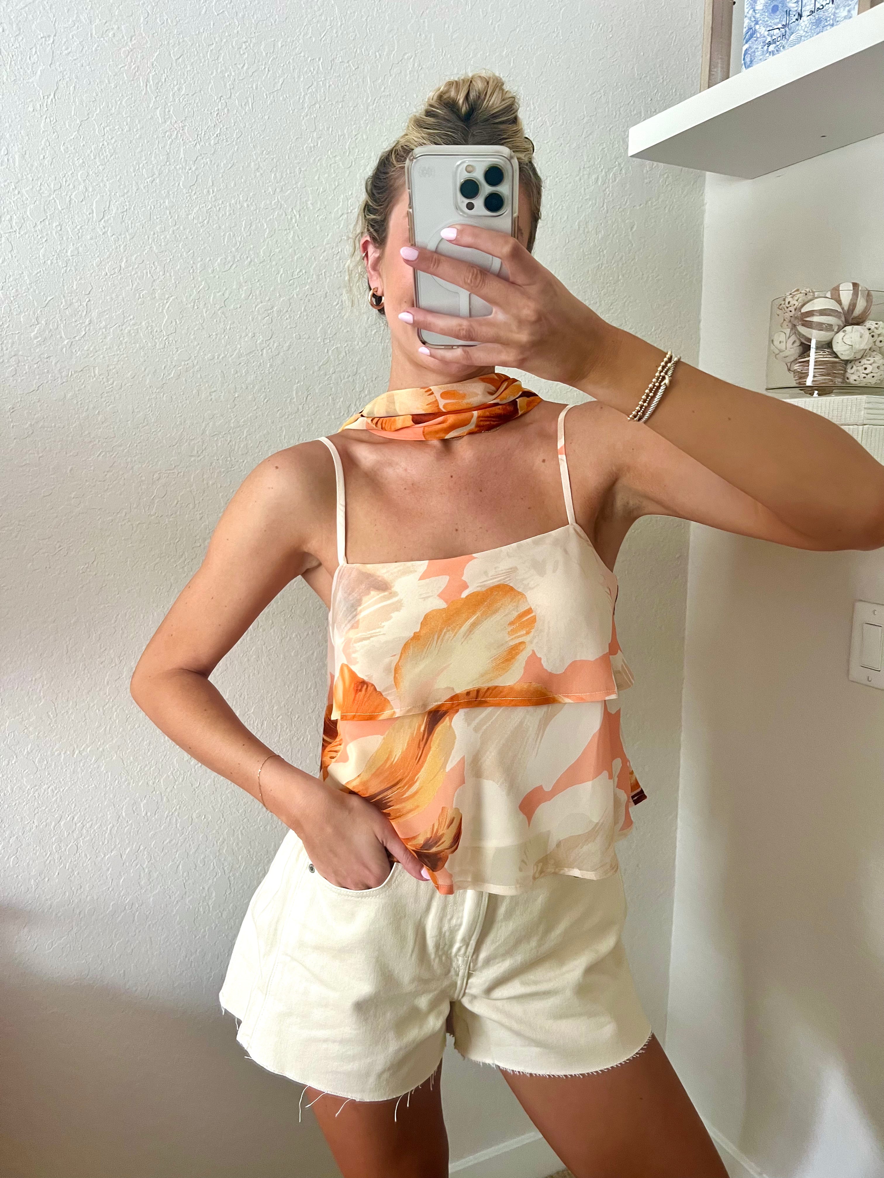 Woman taking a mirror selfie wearing a tank top with orange floral pattern, a neck scarf, and cream shorts.