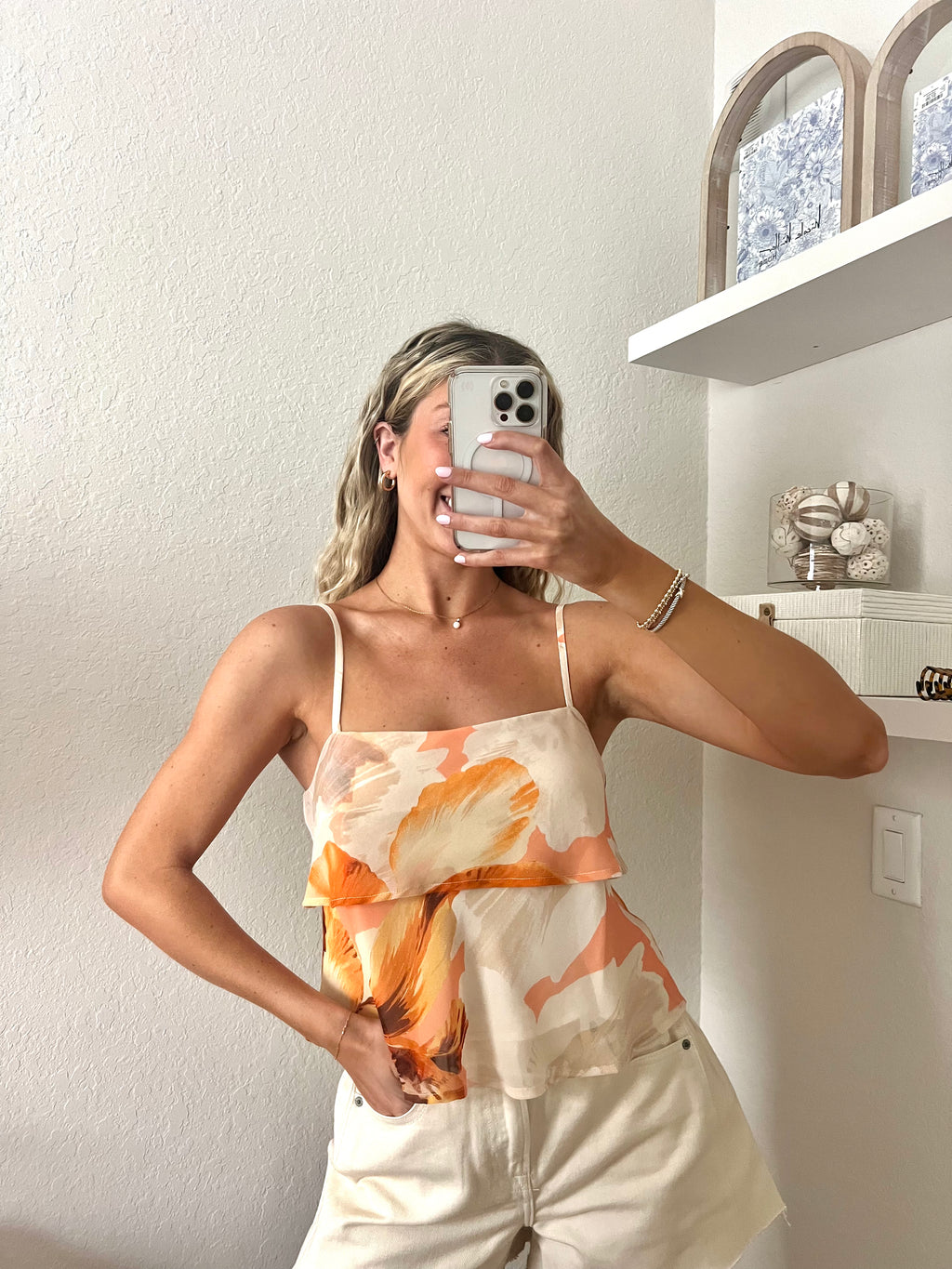 Woman taking a mirror selfie wearing a tank top with orange floral pattern and cream shorts.