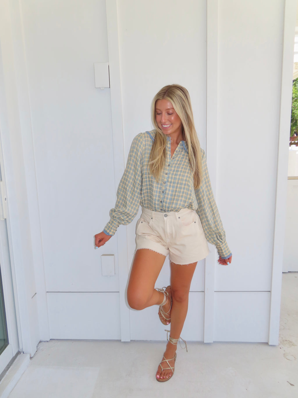 A woman posing in a light green and white checkered long-sleeve button-down shirt, cream-colored denim shorts, and brown lace-up sandals, set against a white door background.