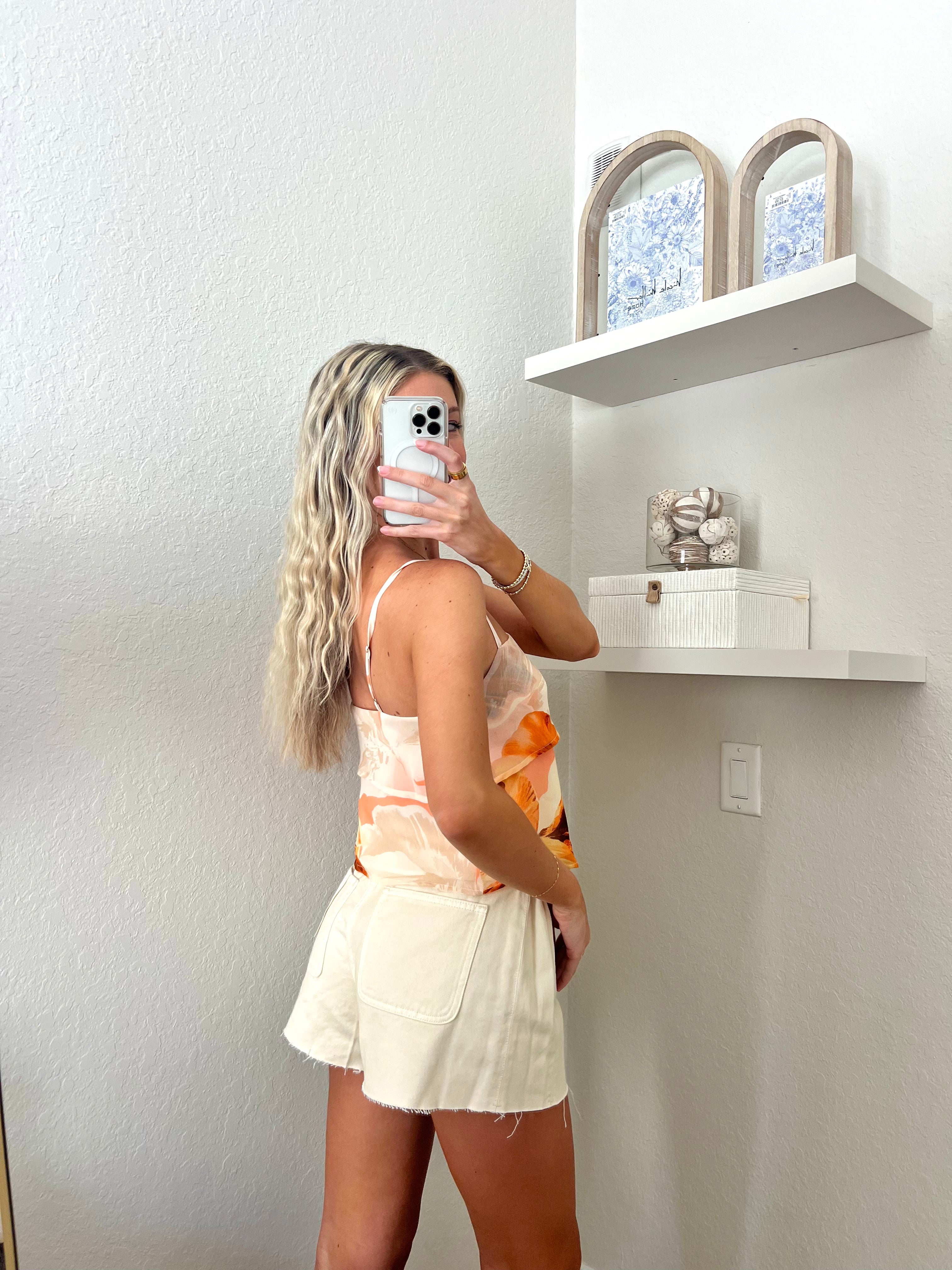 Woman taking a mirror selfie against a plain wall wearing an orange tank and cream denim flare shorts.