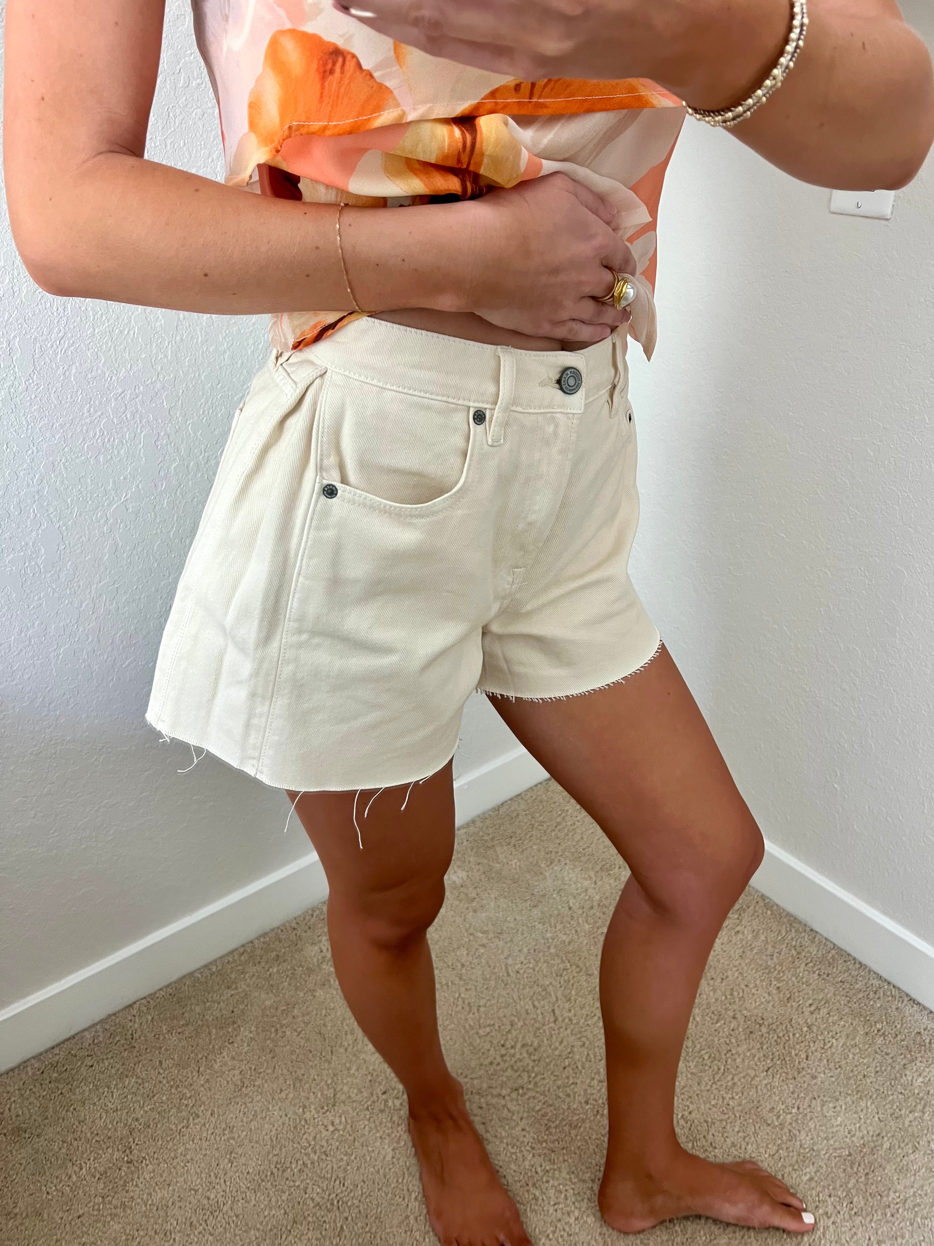 Woman taking a mirror selfie against a plain wall wearing an orange tank and cream denim flare shorts.