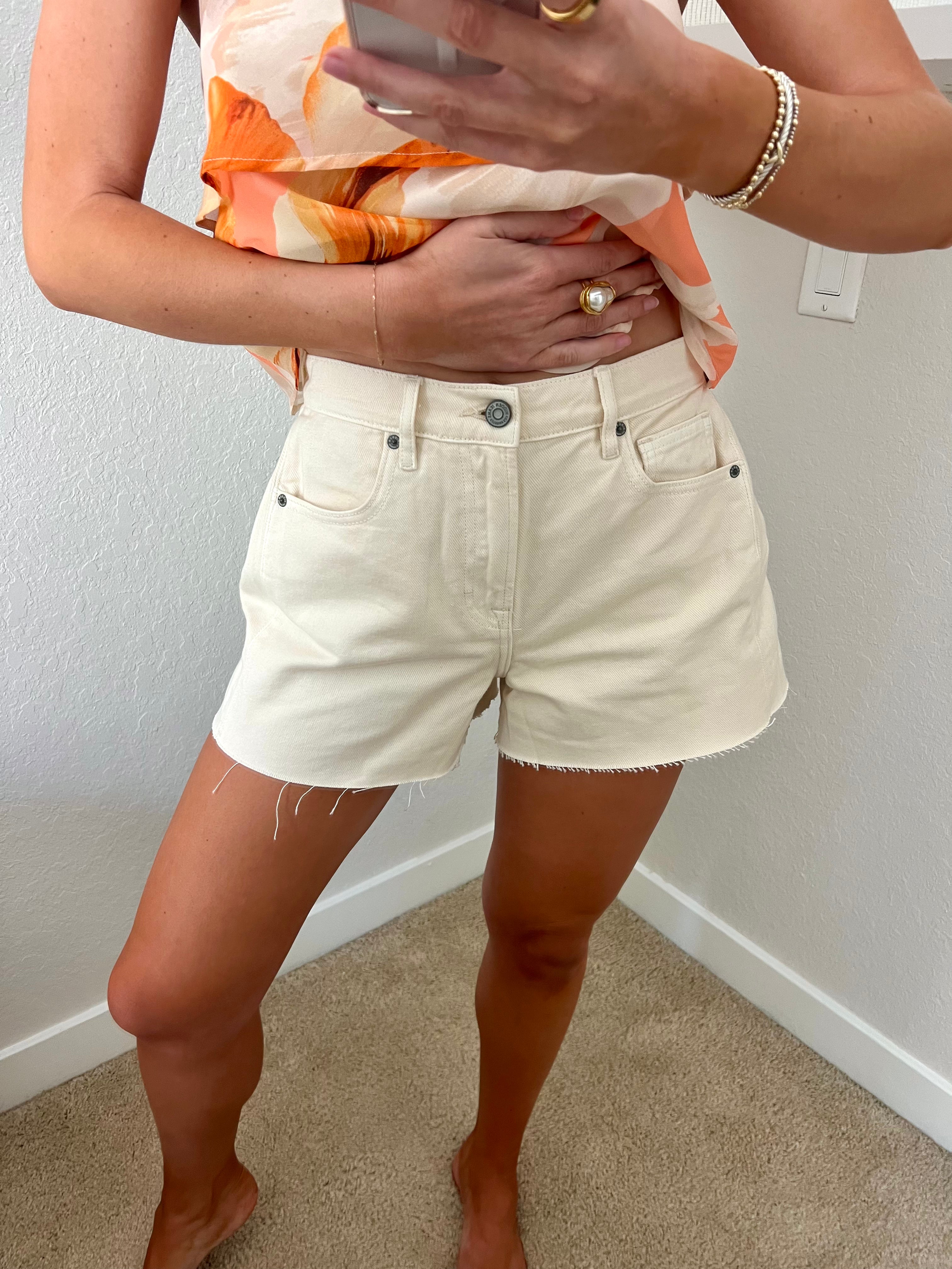 Woman taking a mirror selfie against a plain wall wearing an orange tank and cream denim flare shorts.