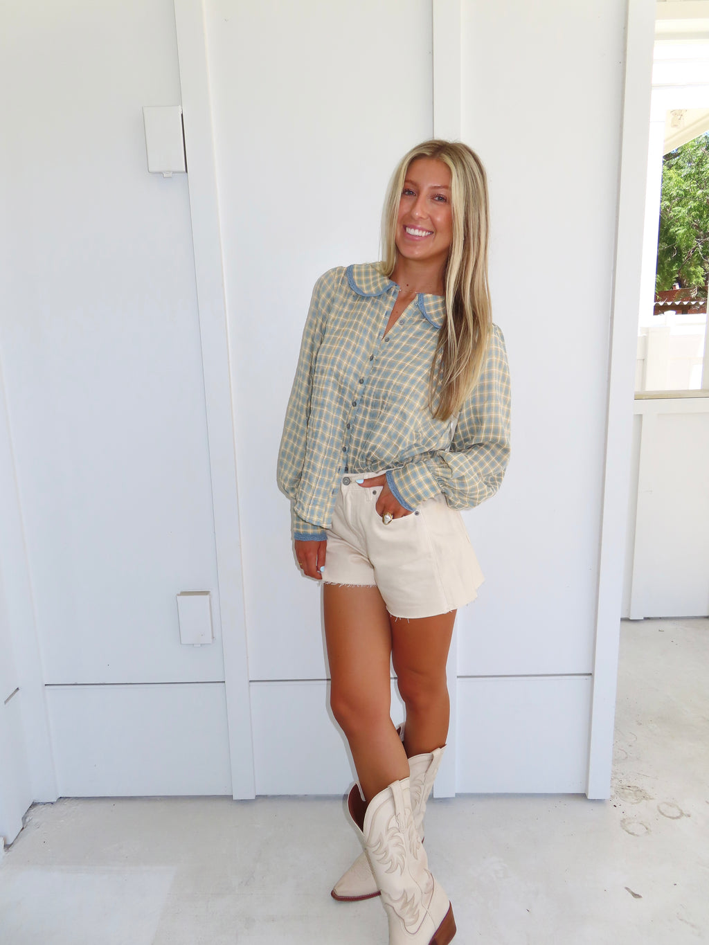 A woman posing against a white wall wearing a blue and cream plaid button-down shirt, cream shorts, and white cowboy boots.