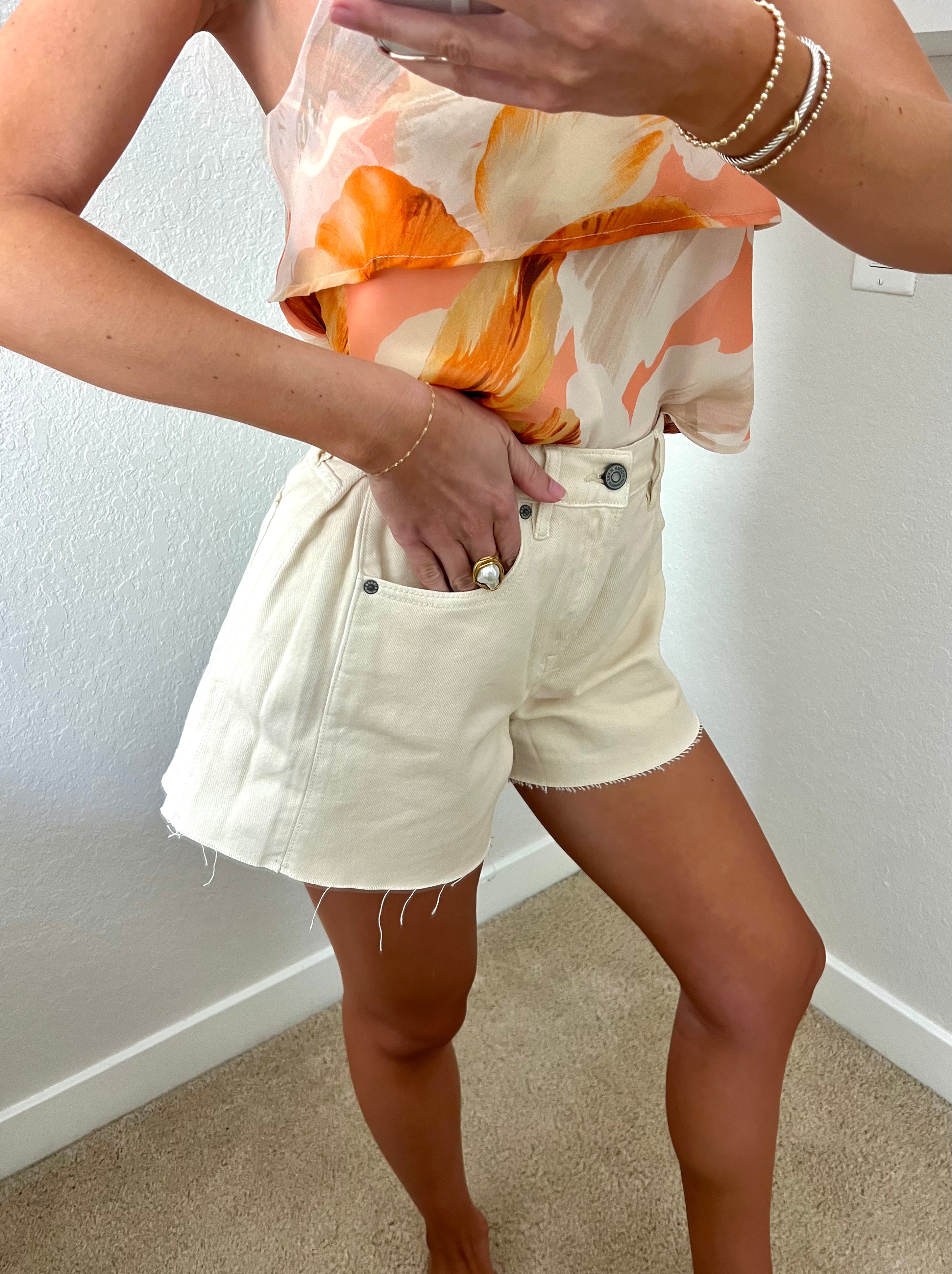 Woman taking a mirror selfie against a plain wall wearing an orange tank and cream denim flare shorts.