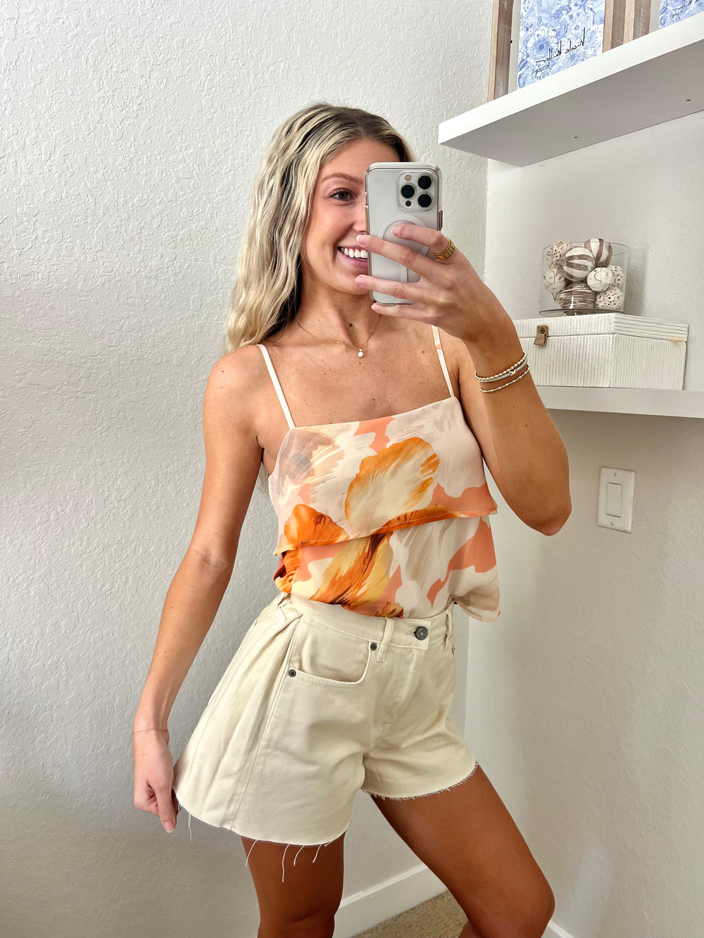 Woman taking a mirror selfie against a plain wall wearing an orange tank and cream denim flare shorts.