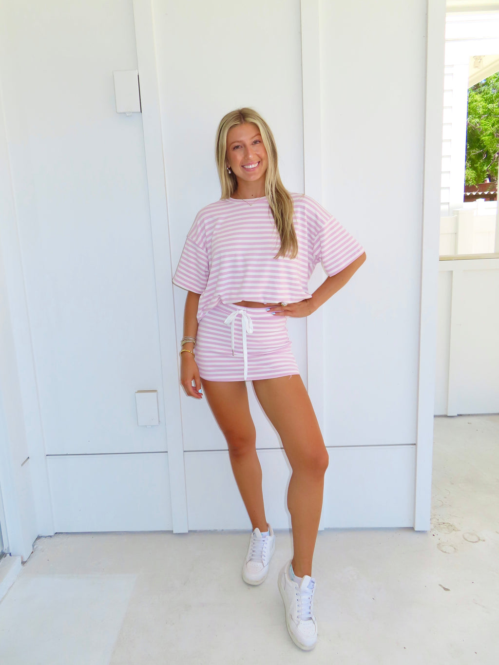 Woman wearing a pink and white striped skort set with white sneakers in front of a white wall.