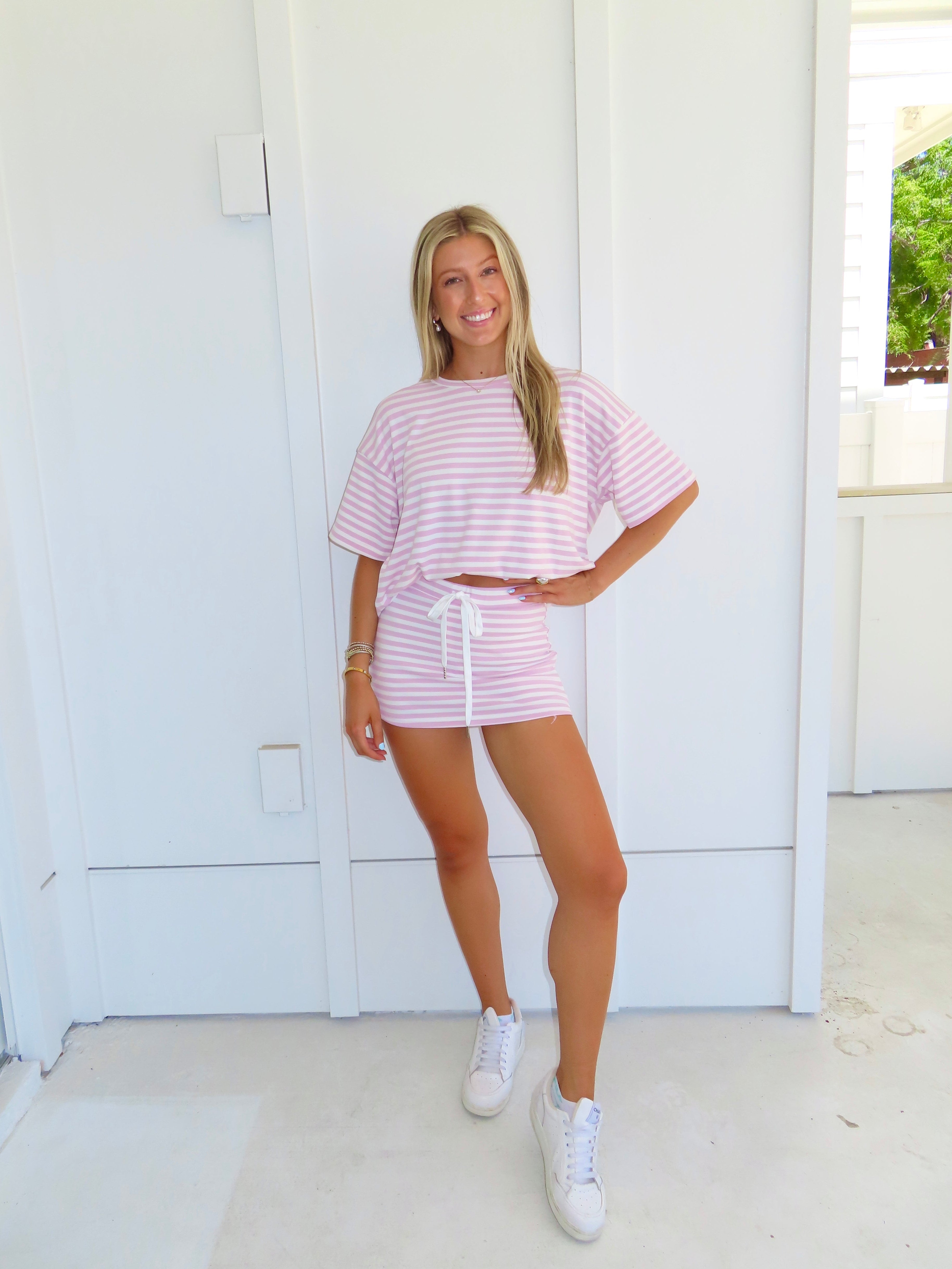 Woman wearing a pink and white striped skort set with white sneakers in front of a white wall.
