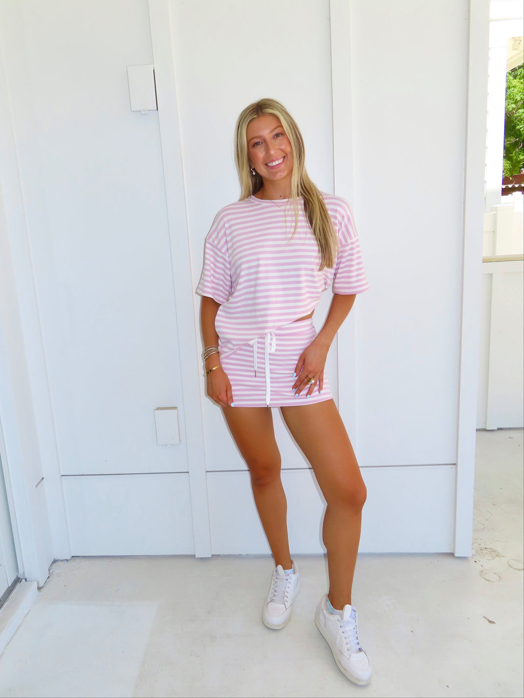 Woman wearing a pink and white striped skort set with white sneakers in front of a white wall.