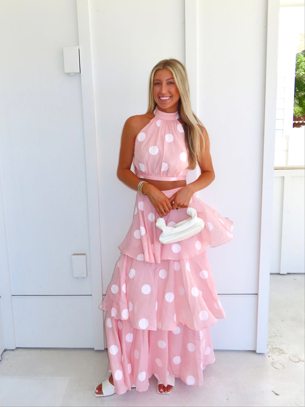 Woman wearing a pink polka dot skirt set holding a white clutch on a bright white patio.