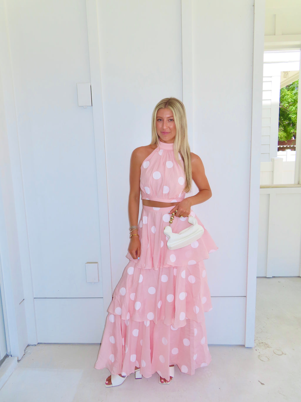 Woman wearing a pink polka dot skirt set holding a white clutch on a bright white patio.